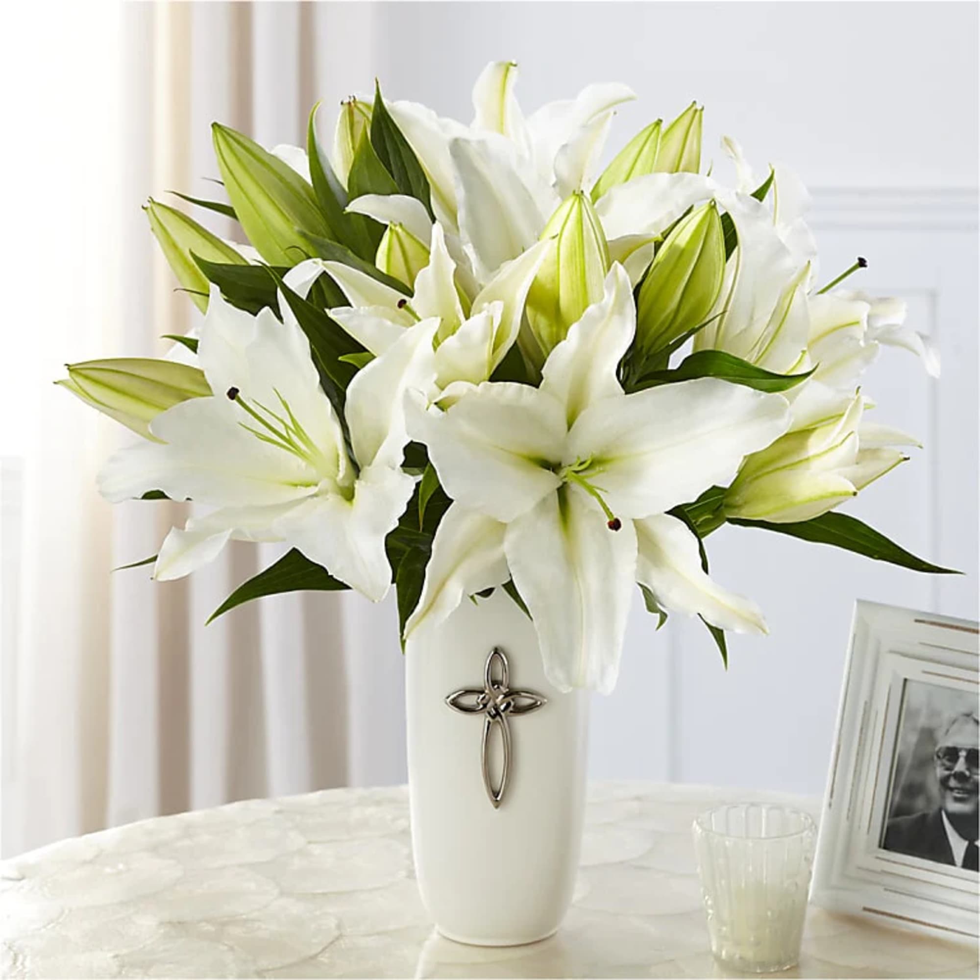 White lilies in a tall white vase with a silver cross emblem on a table.