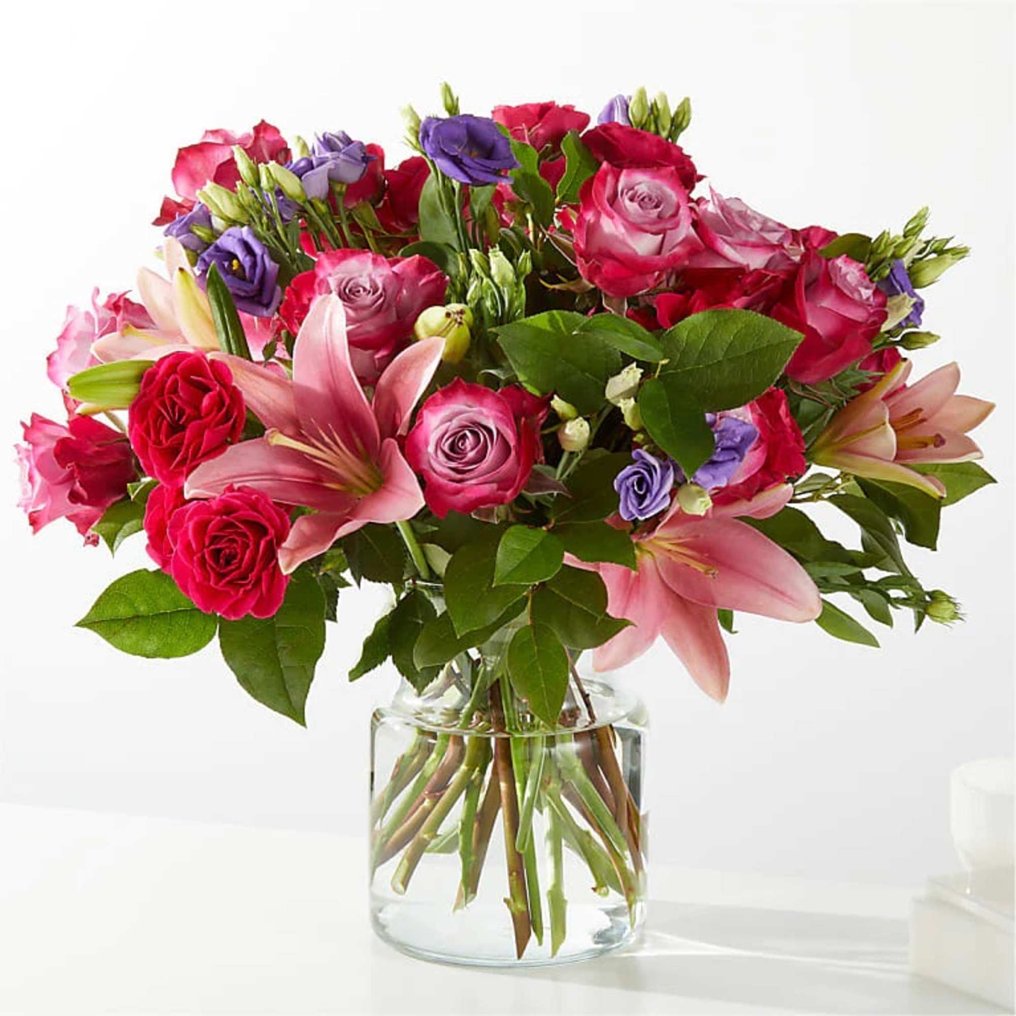 Mixed bouquet of pink lilies, red and pink roses, and purple blooms in a clear glass vase