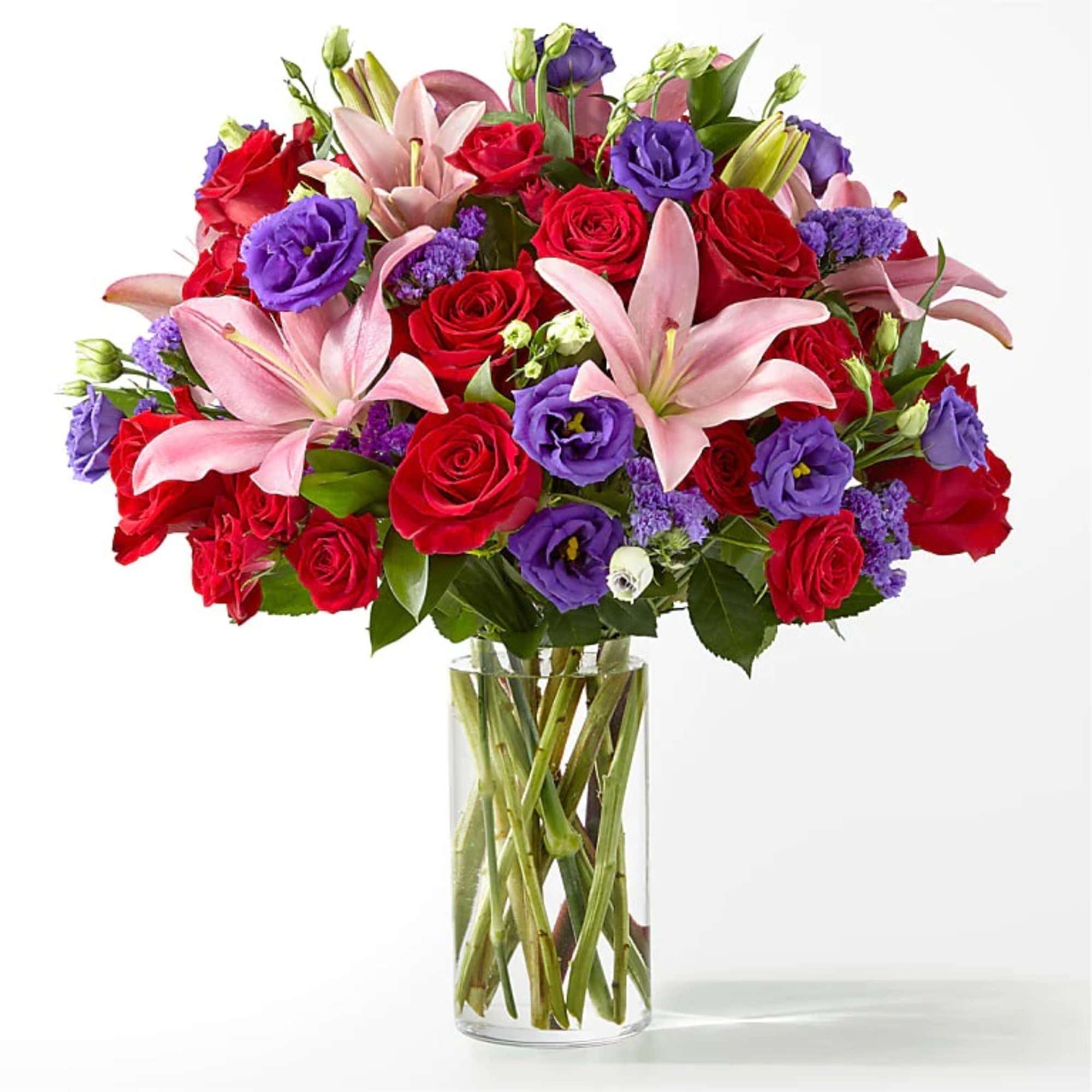 Tall glass vase of pink lilies, red roses, and purple blooms in a full mixed flower arrangement