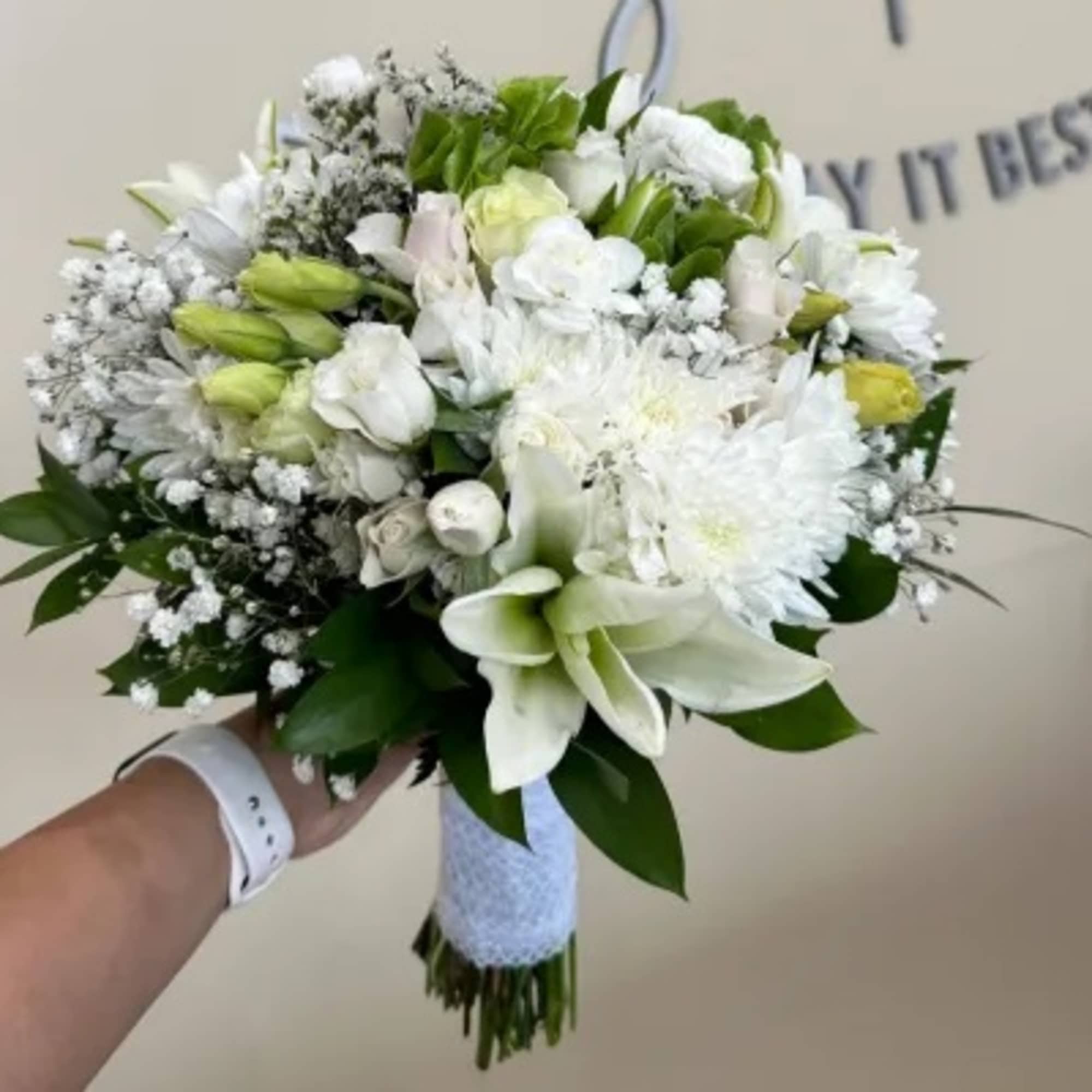 Hand-tied white bouquet with lilies, roses, and mums wrapped in white ribbon, held against a neutral background.