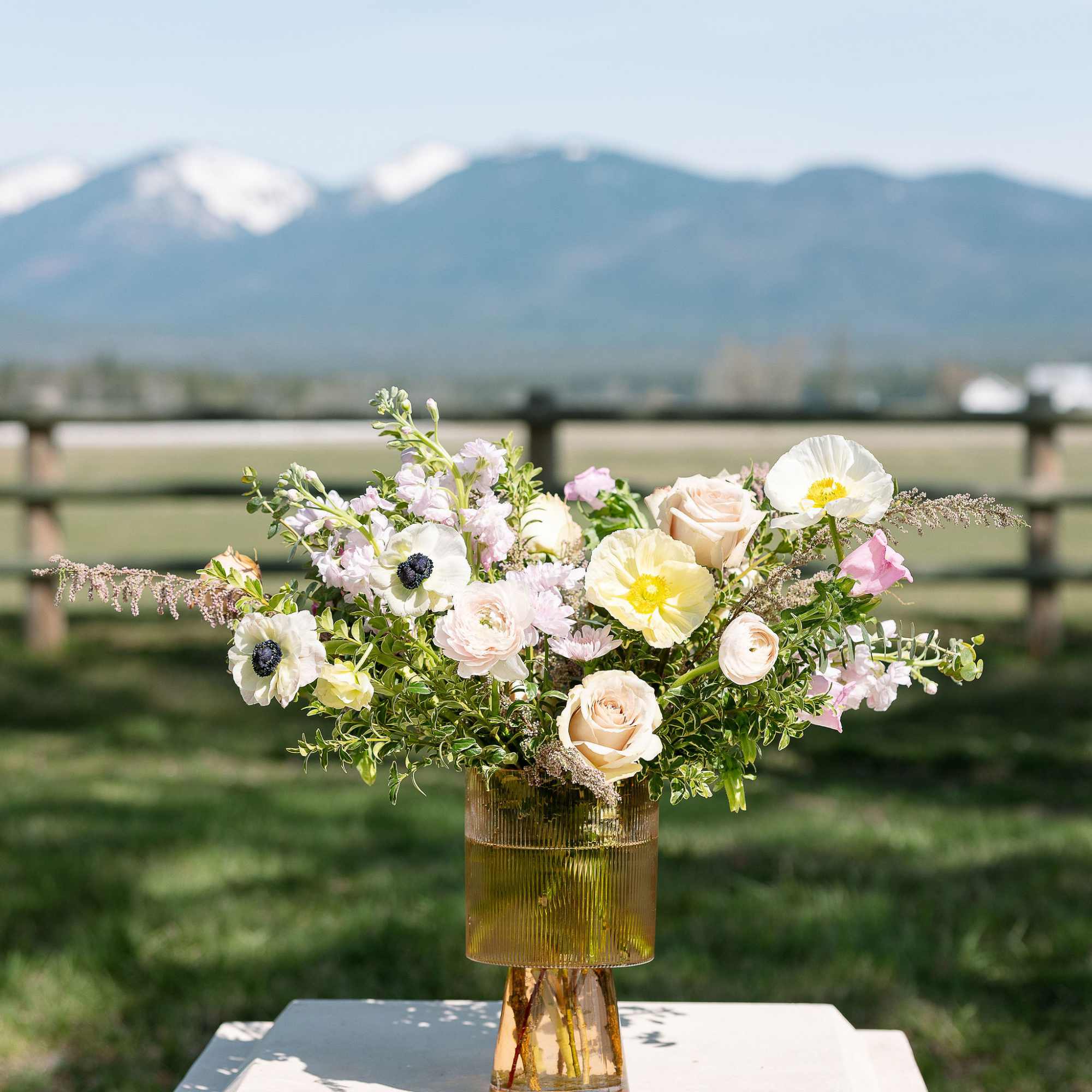 Garden-style arrangement of white and pastel flowers in an amber glass vase set outdoors with mountains behind.