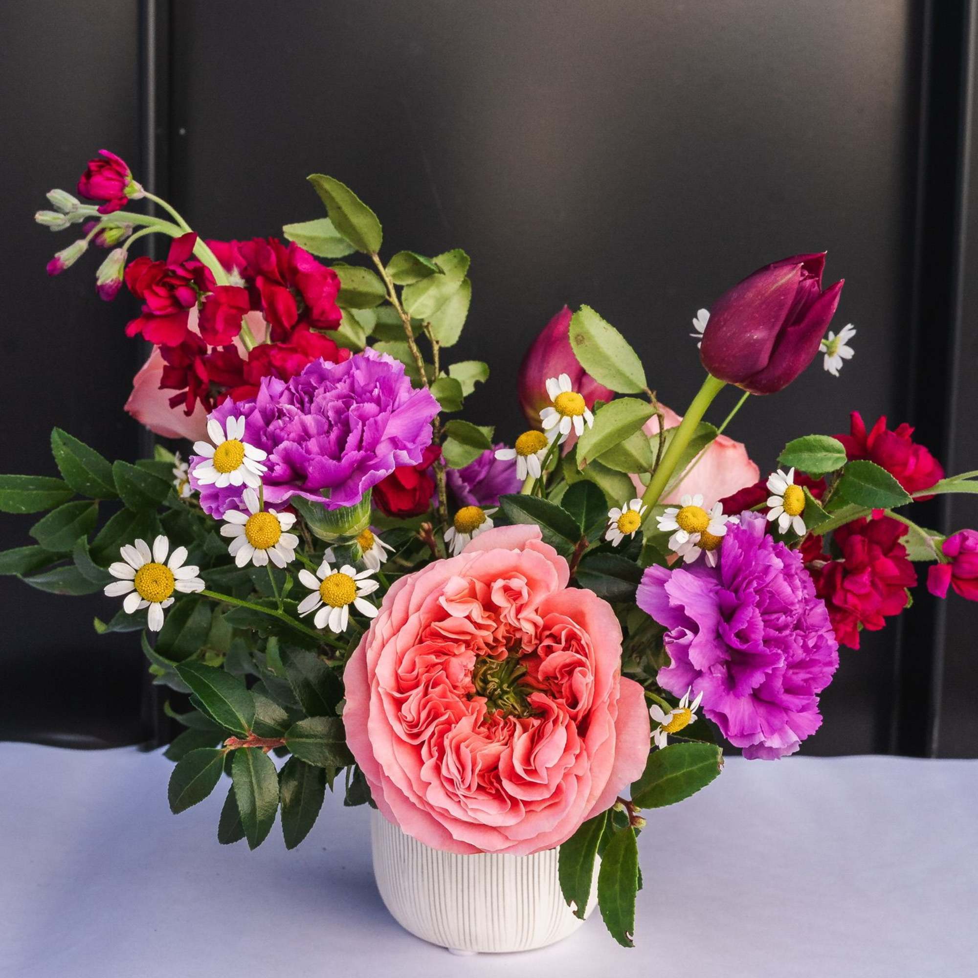 Compact arrangement of pink roses, purple carnations, red stock, and small white daisies in a white ceramic vase.
