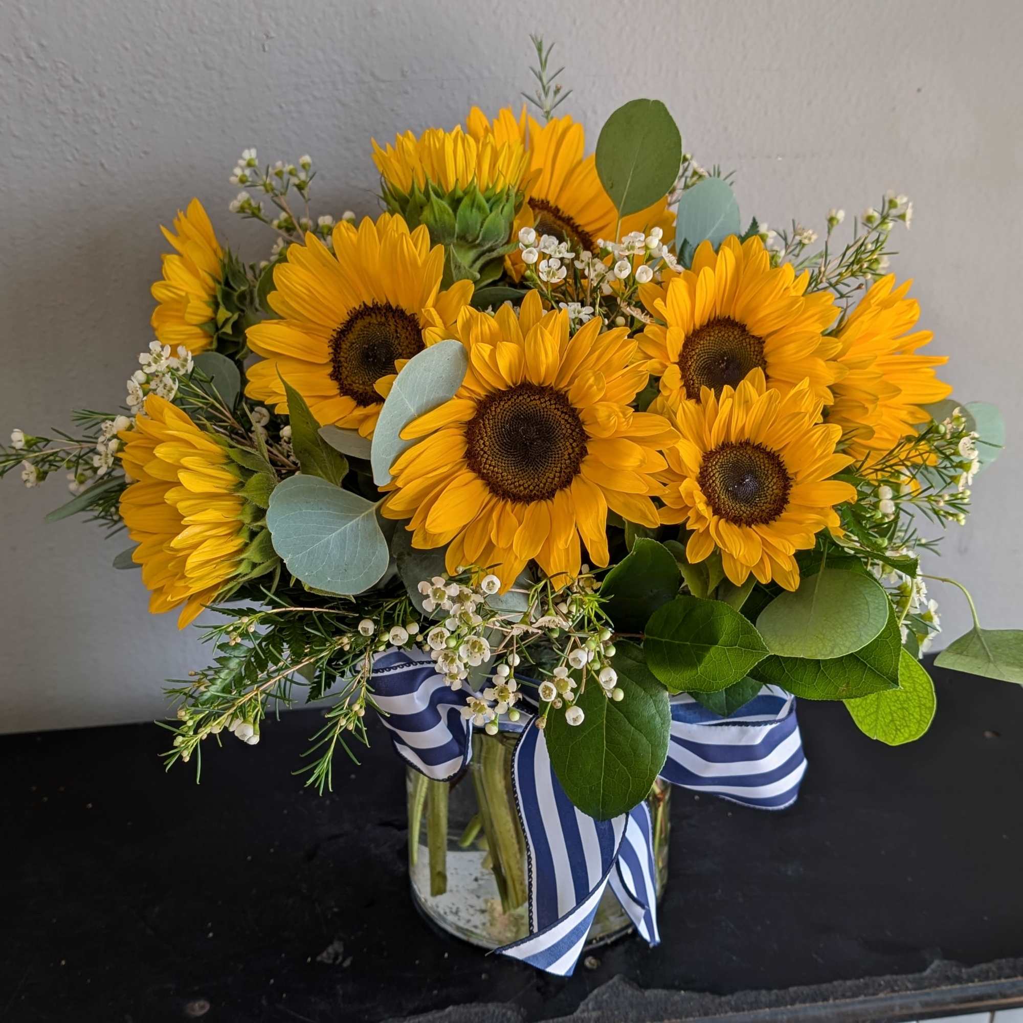 Arrangement of bright yellow sunflowers with small white blooms in a clear glass vase tied with a blue striped ribbon.