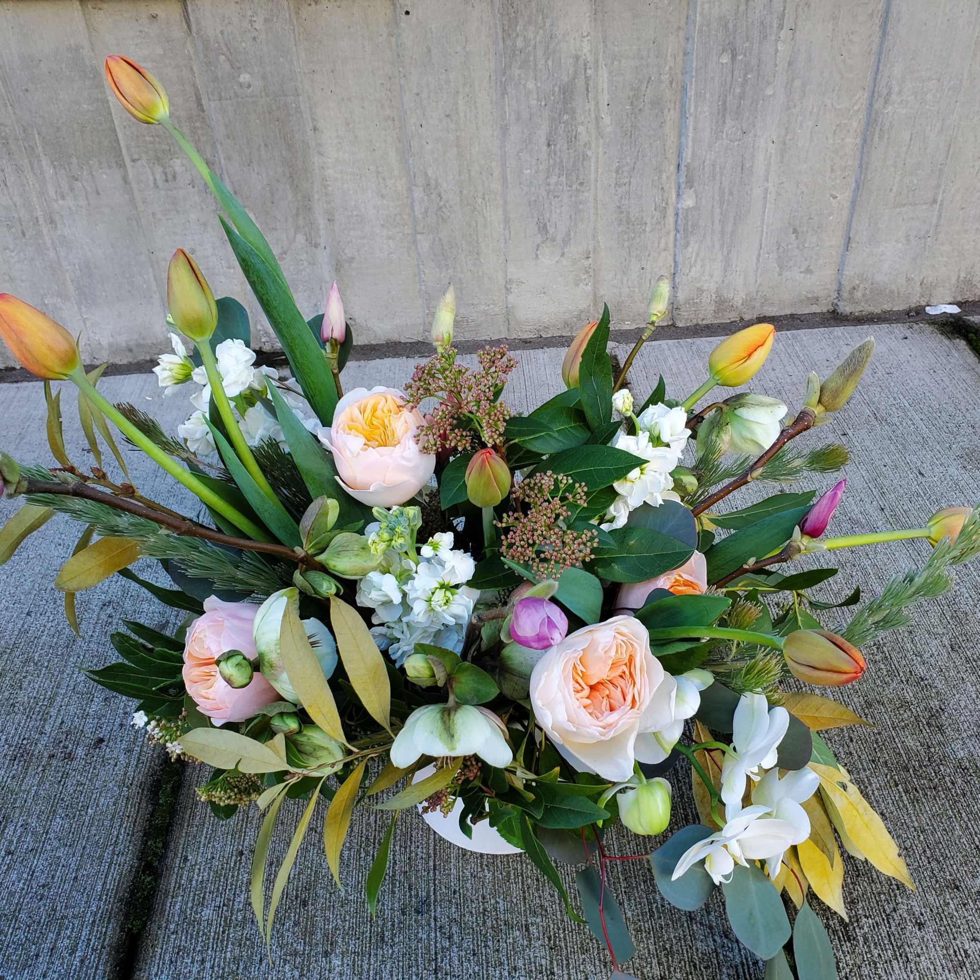 Low arrangement of peach roses, tulips, and white orchids in a white bowl on concrete