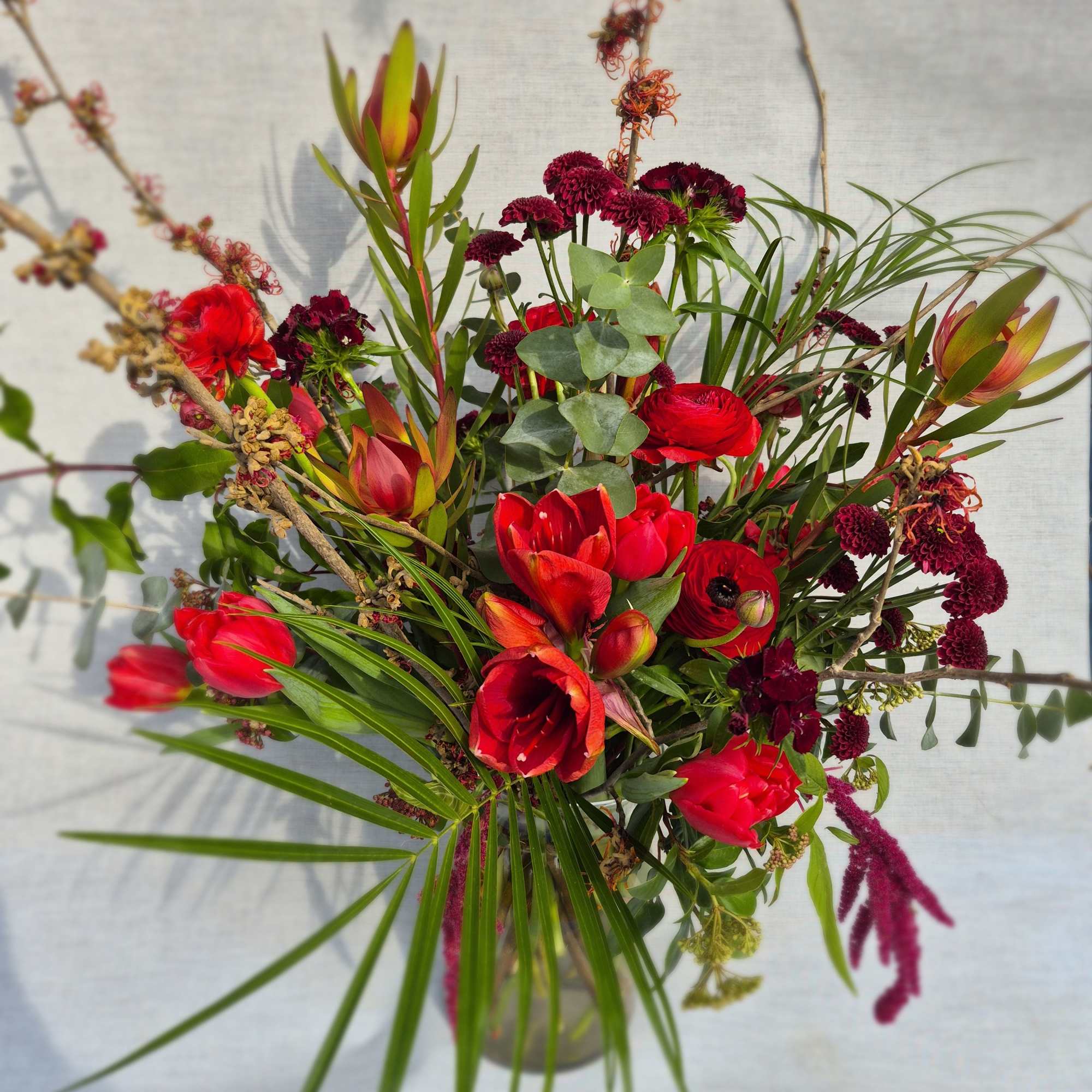 Tall arrangement of red tulips, ranunculus, and dark blooms with tropical foliage in a clear glass vase