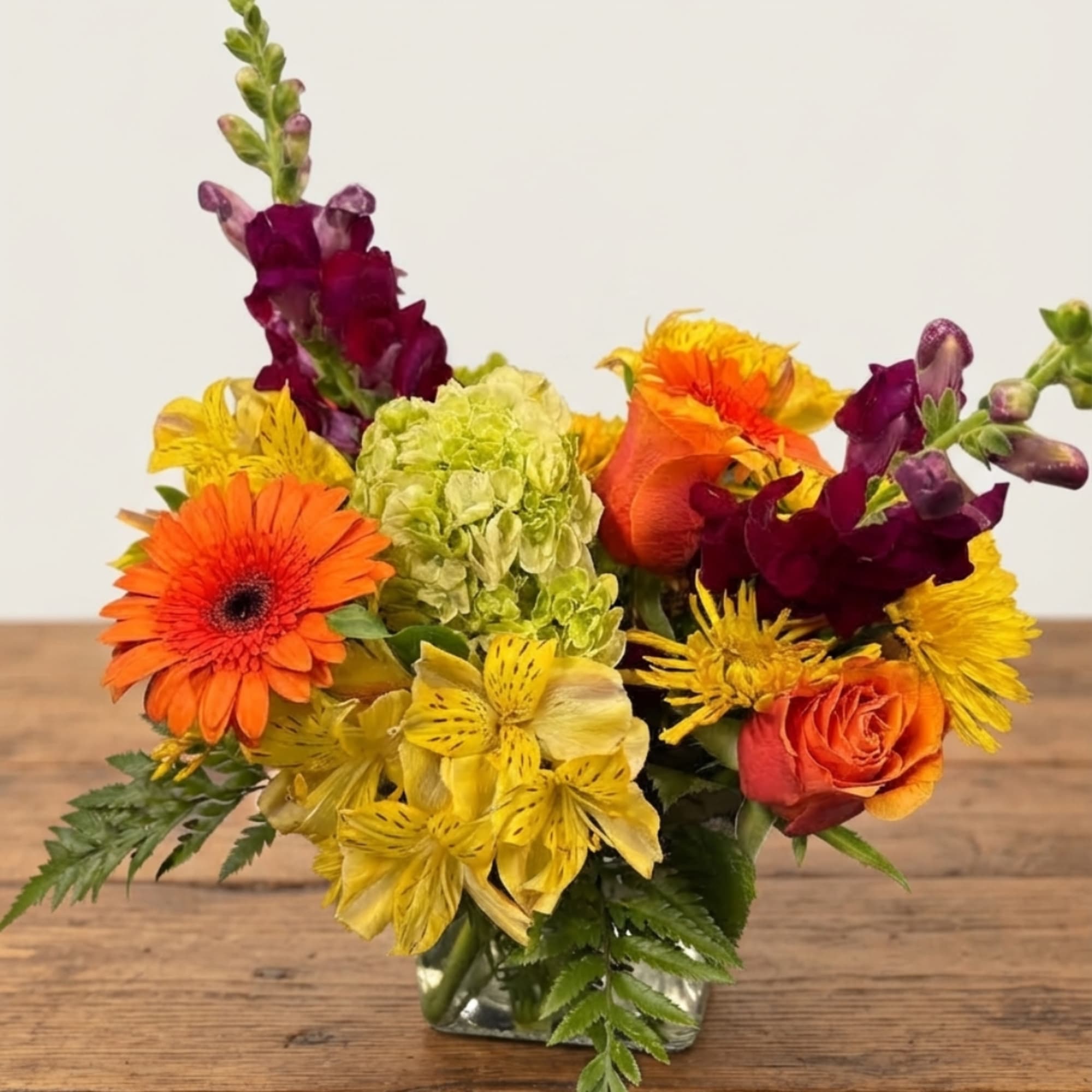 Colorful bouquet with orange gerbera daisies, yellow blooms, and purple snapdragons in a glass vase
