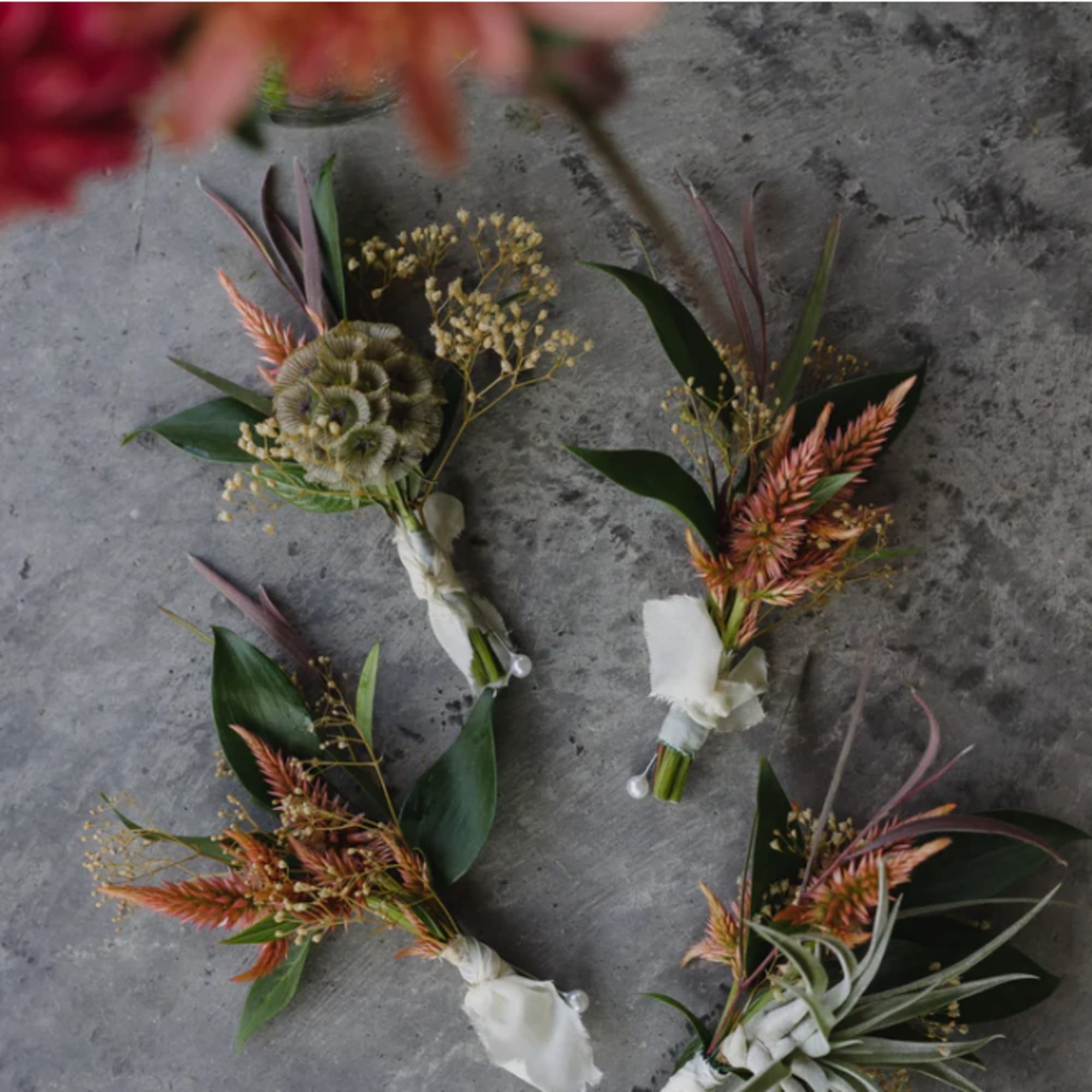 Four small floral boutonnieres with orange plumes and greenery on a gray surface