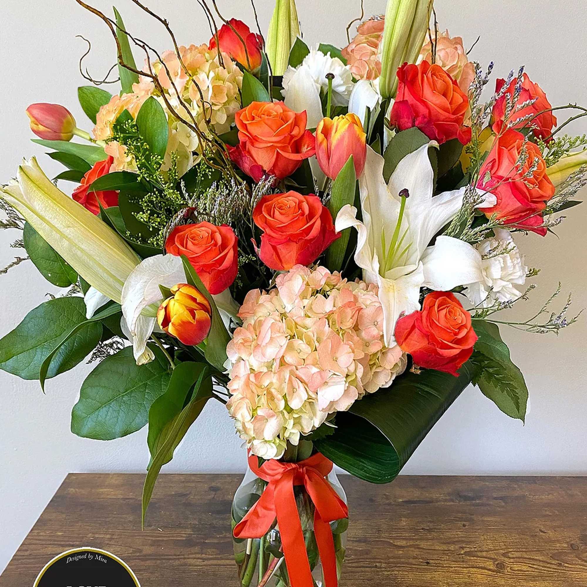 Tall mixed arrangement of orange roses, tulips, white lilies, and peach hydrangeas in a clear vase with orange ribbon
