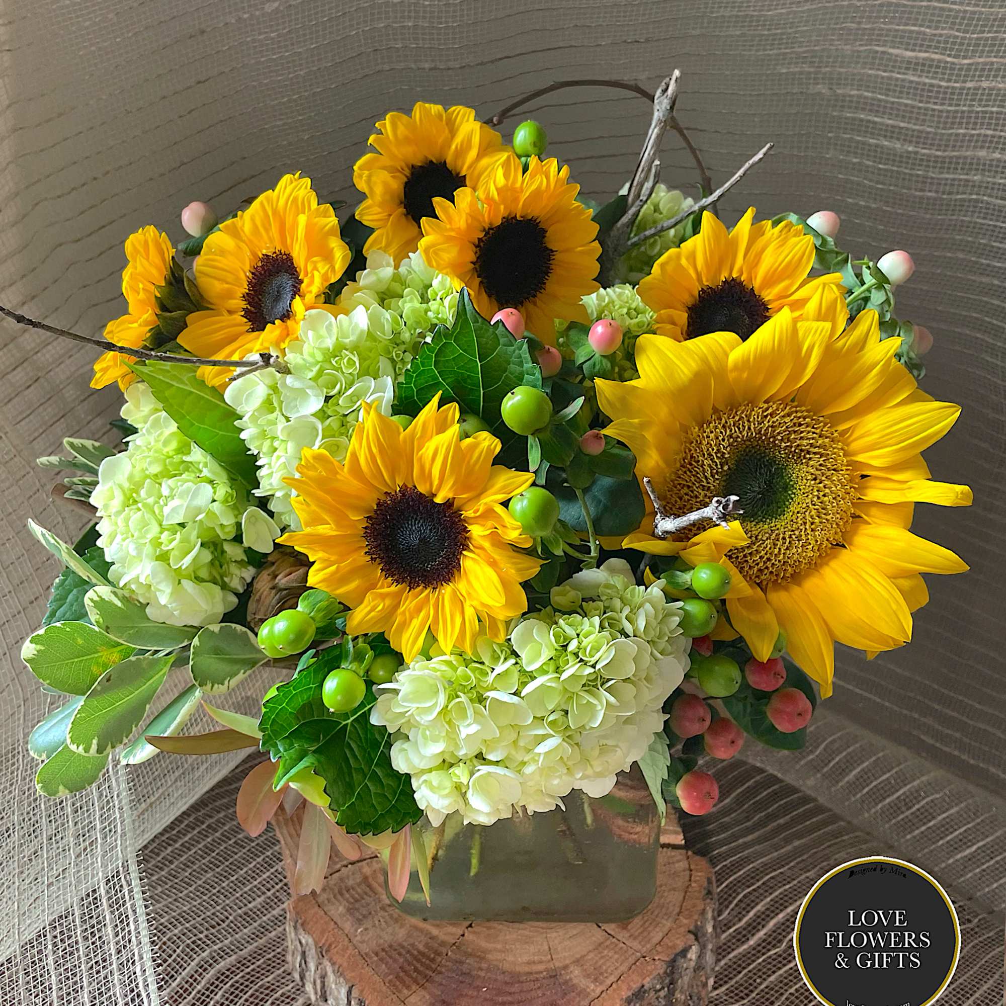 Arrangement of yellow sunflowers and white hydrangeas with berries in a square glass vase