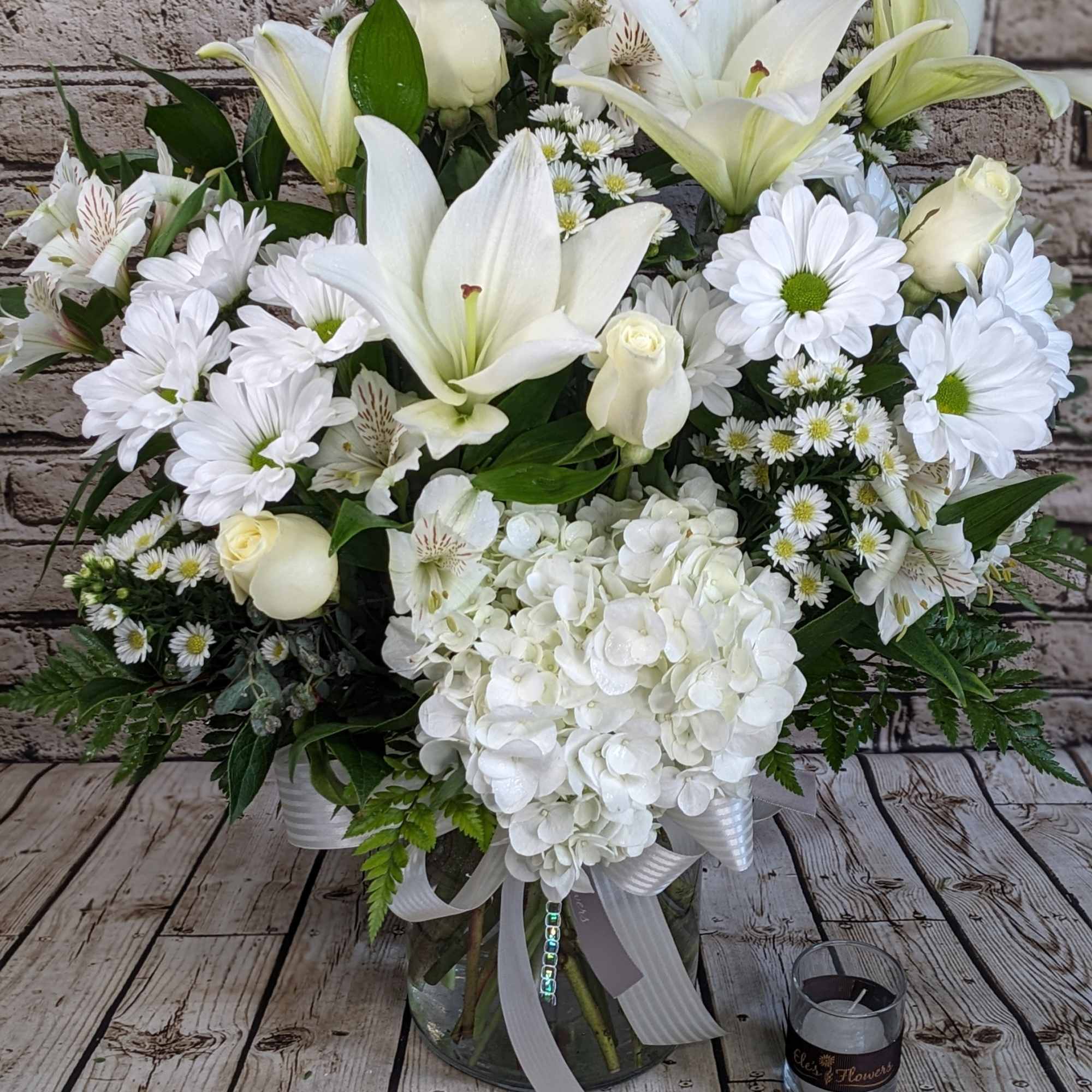 All-white arrangement of lilies, roses, daisies, and hydrangea in a clear glass vase with ribbon.