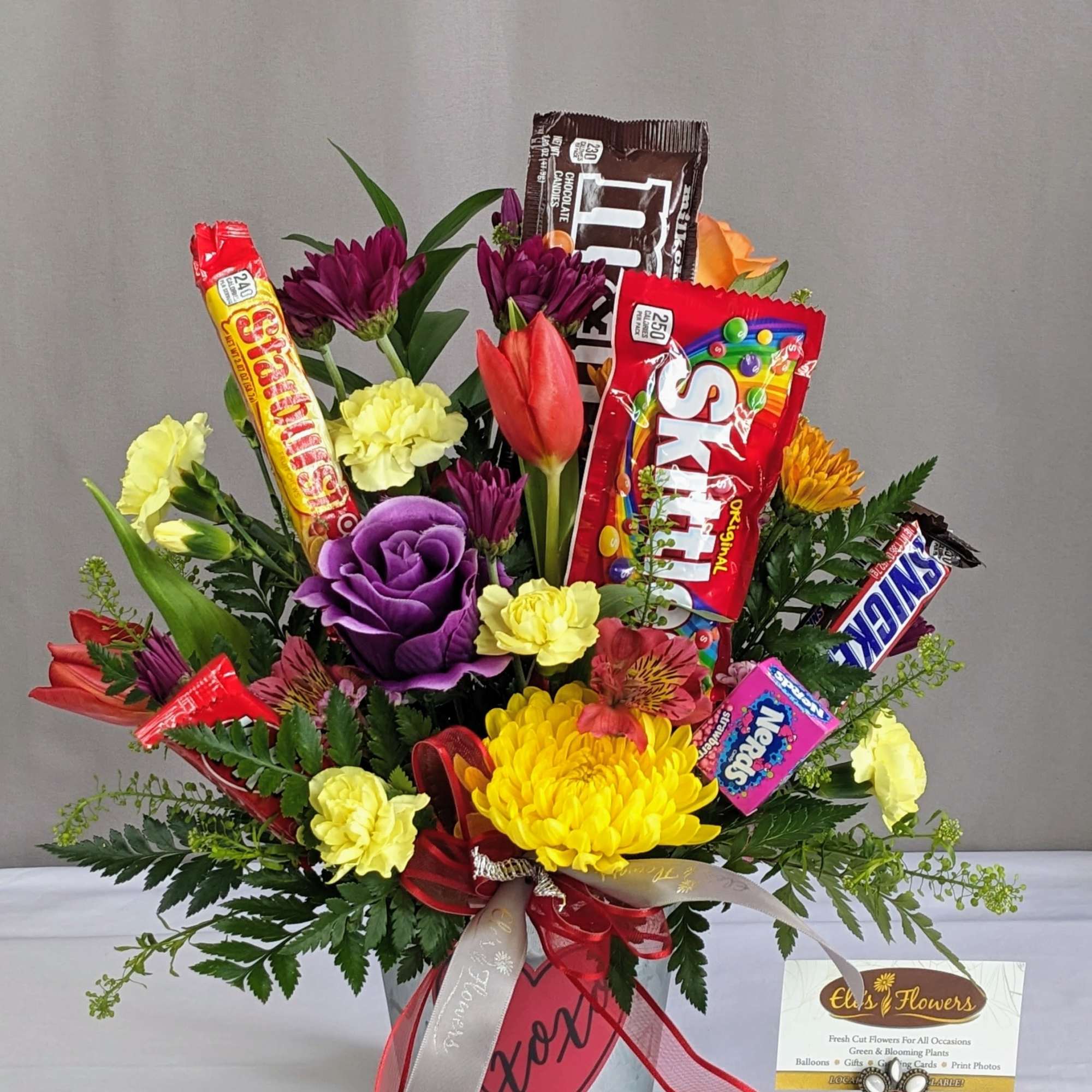 Arrangement of yellow and purple flowers in a metal bucket with assorted candy bars among the blooms.