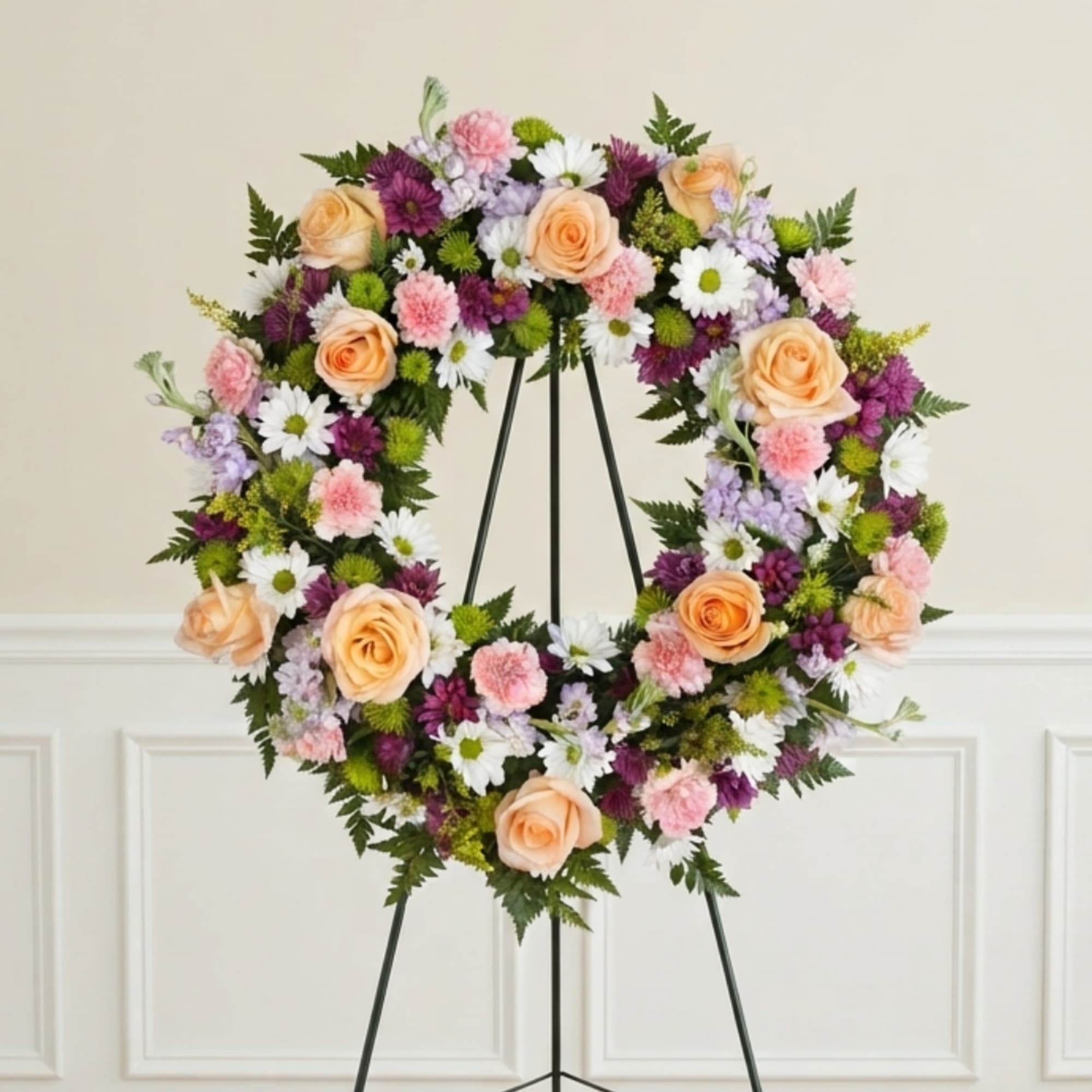 Floral wreath on a black stand with peach roses, daisies, and pink blooms