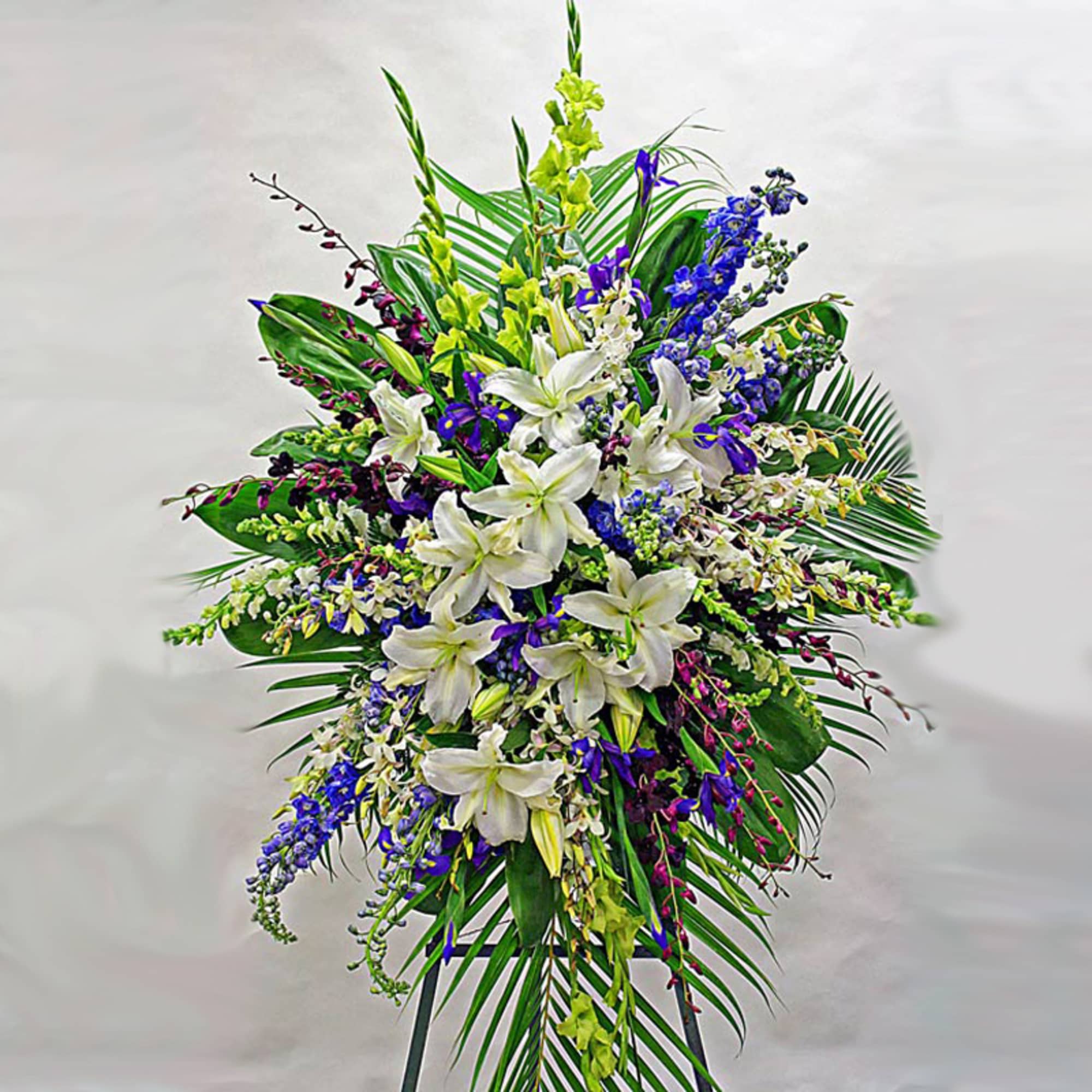 Tall standing spray of white lilies with blue and yellow flowers on palm foliage easel