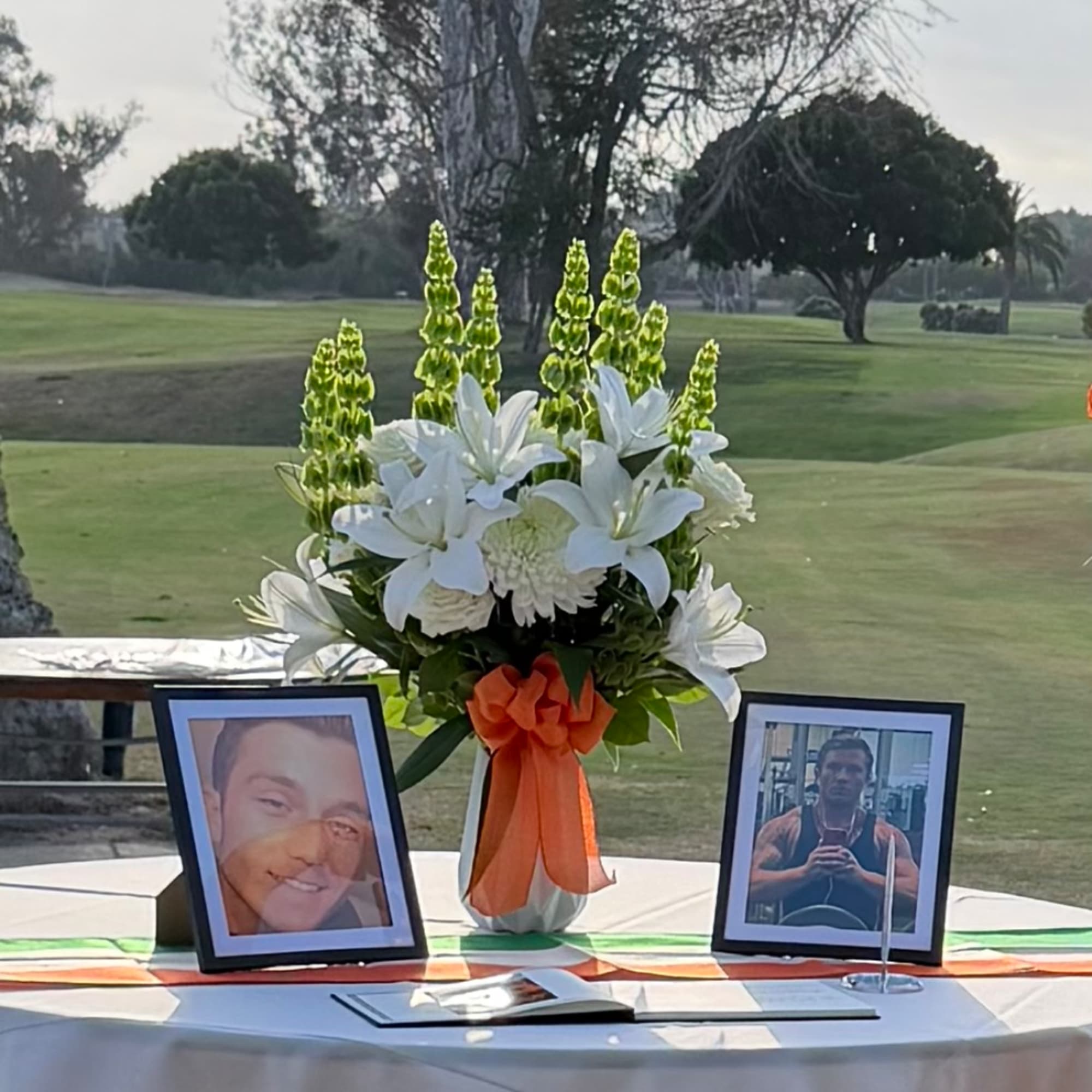 White lilies and green spikes in a vase with orange ribbon, flanked by framed photos