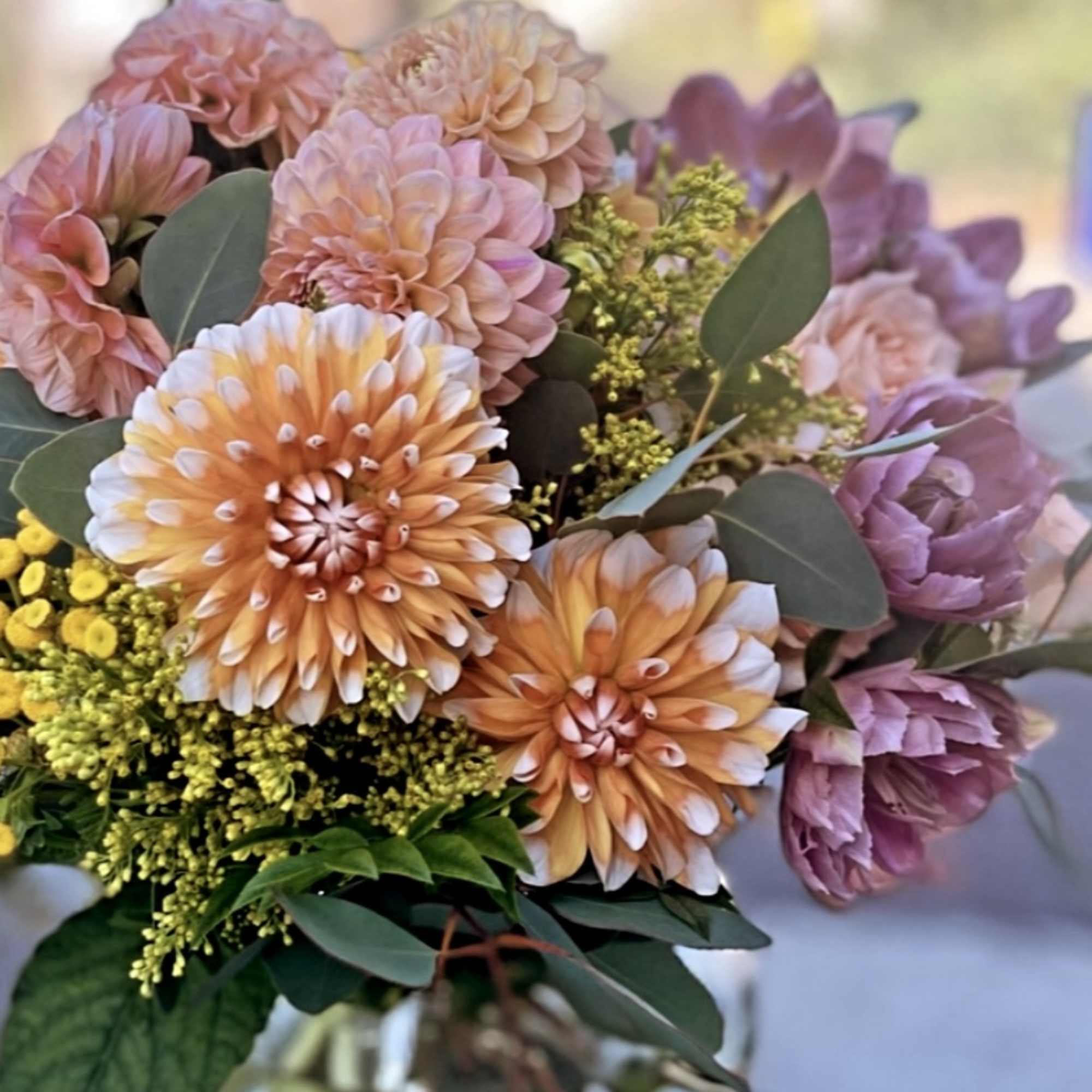 Peach dahlias and mauve blooms arranged with yellow accents in a clear glass vase.
