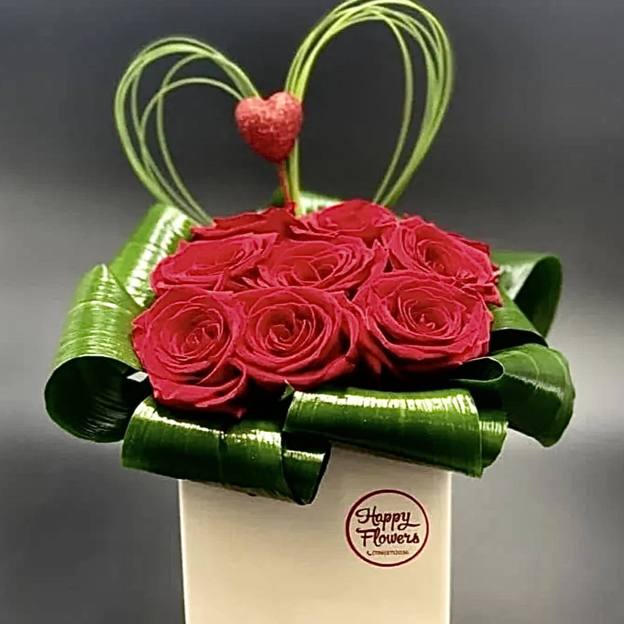 Red roses in a white box with green ribbon loops and a heart topper