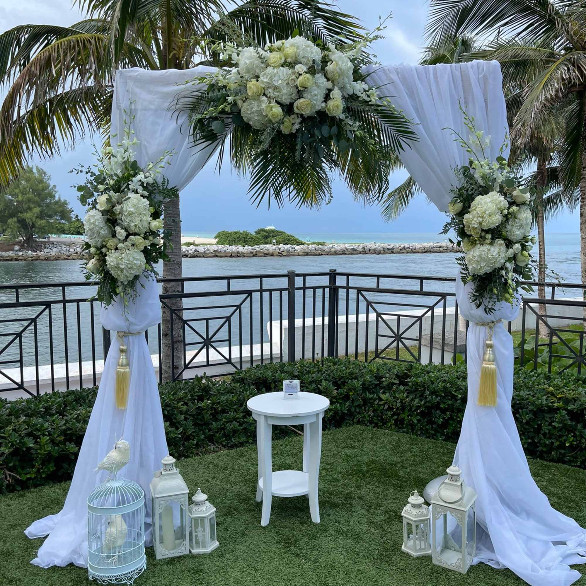 White floral wedding arch with draped fabric and lanterns by the water