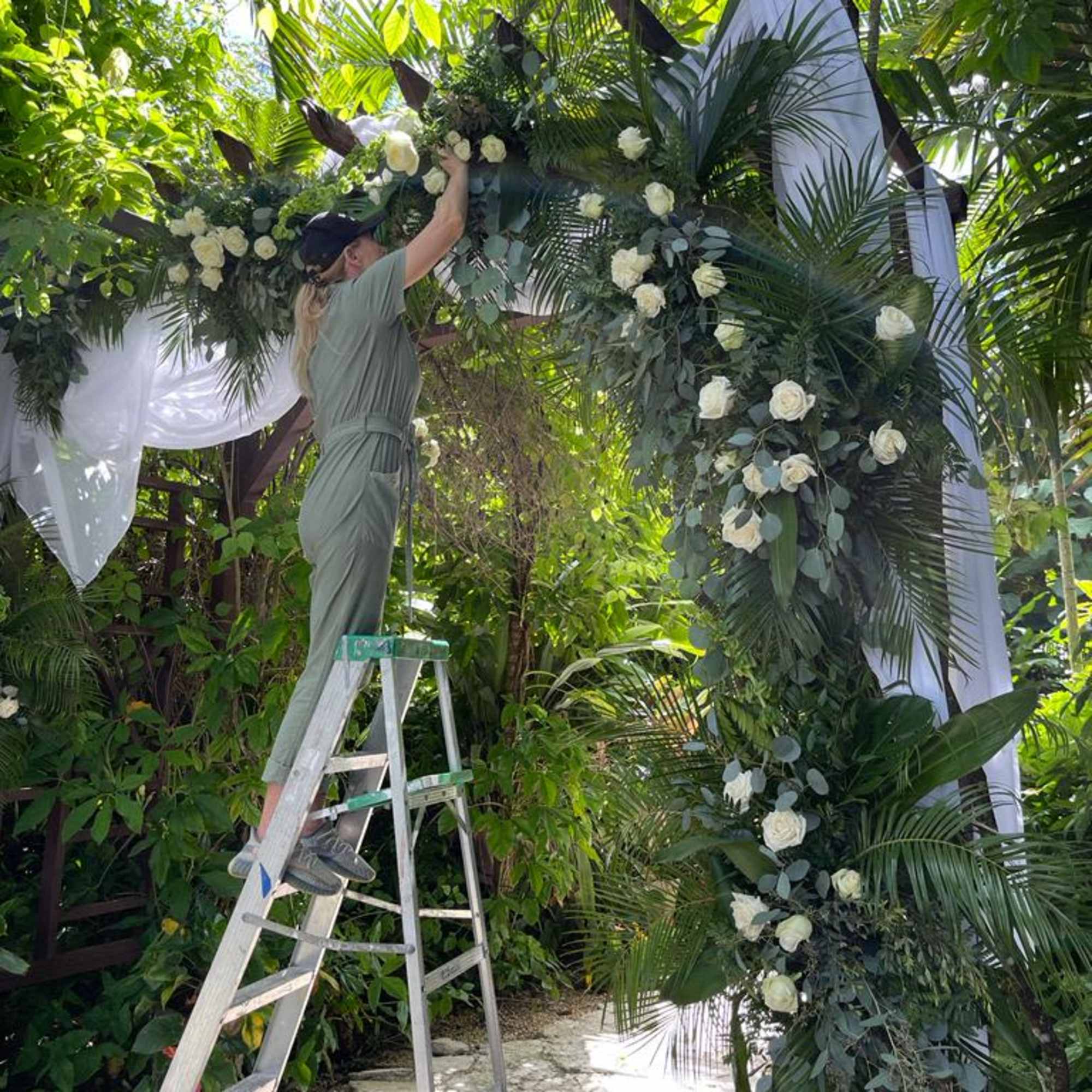 White rose floral arch being decorated outdoors with a ladder