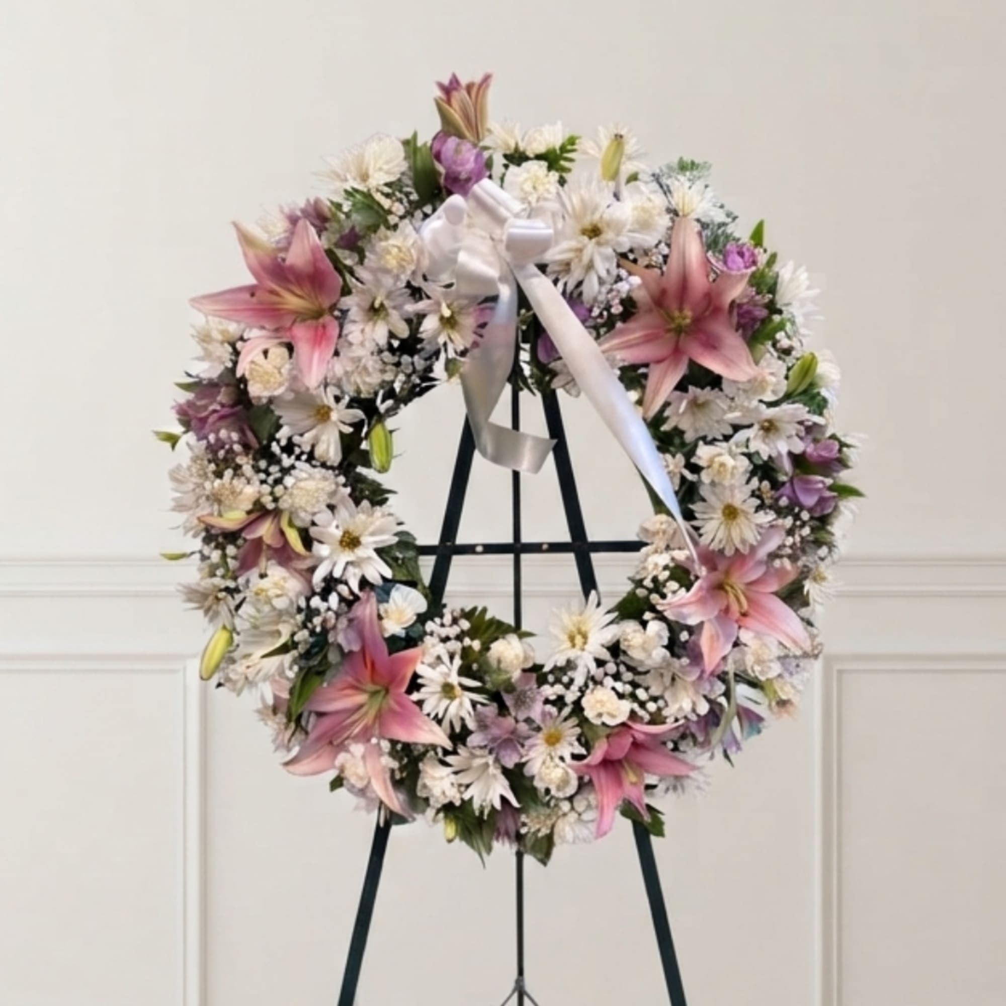 Pink lilies and white daisies arranged in a wreath on a stand with a white ribbon