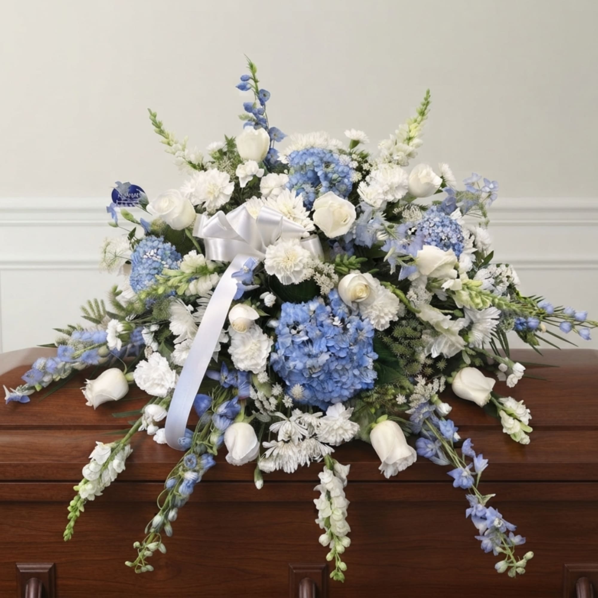 Blue and white funeral spray with roses, hydrangeas, and ribbon on a casket