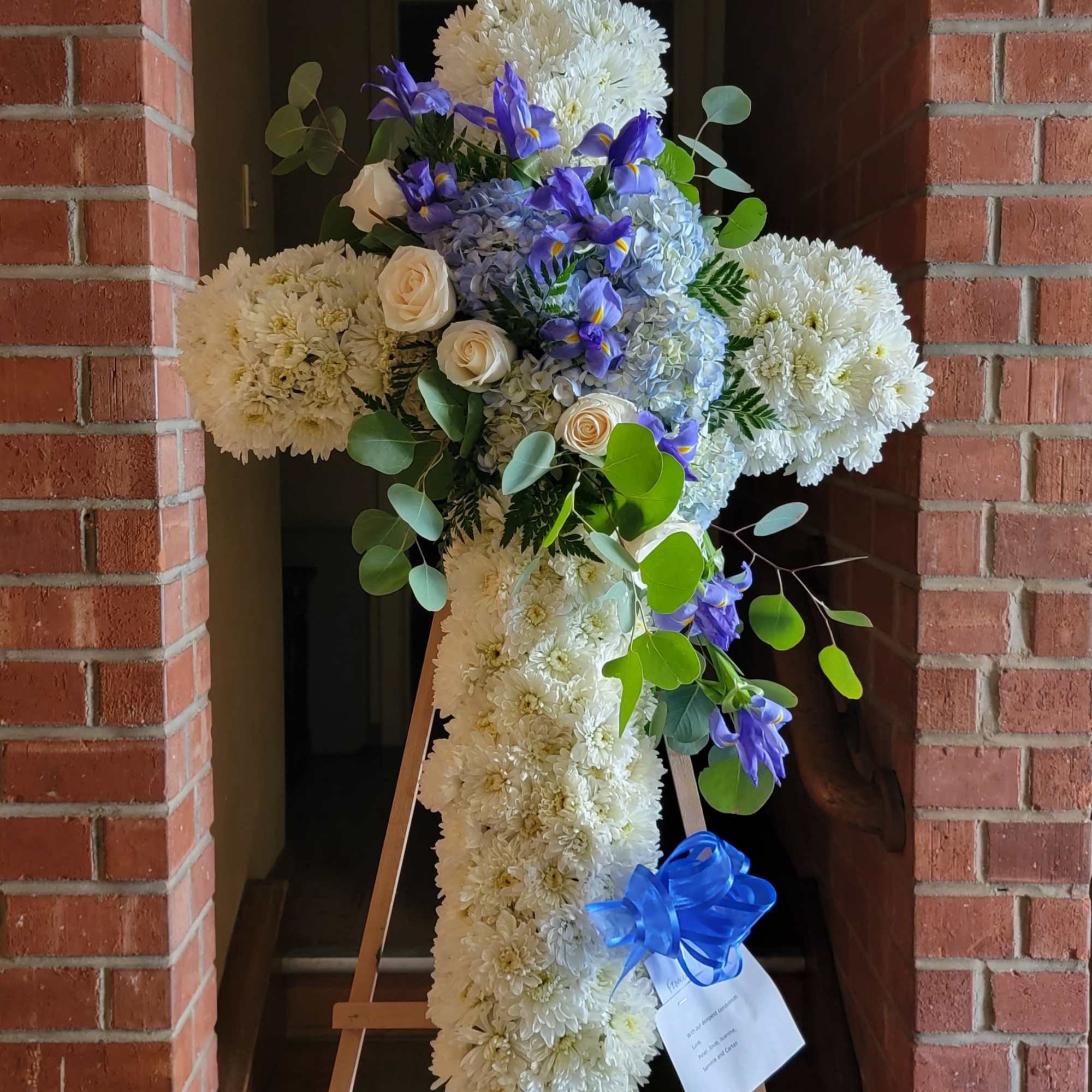 White floral cross on an easel with blue flowers and greenery