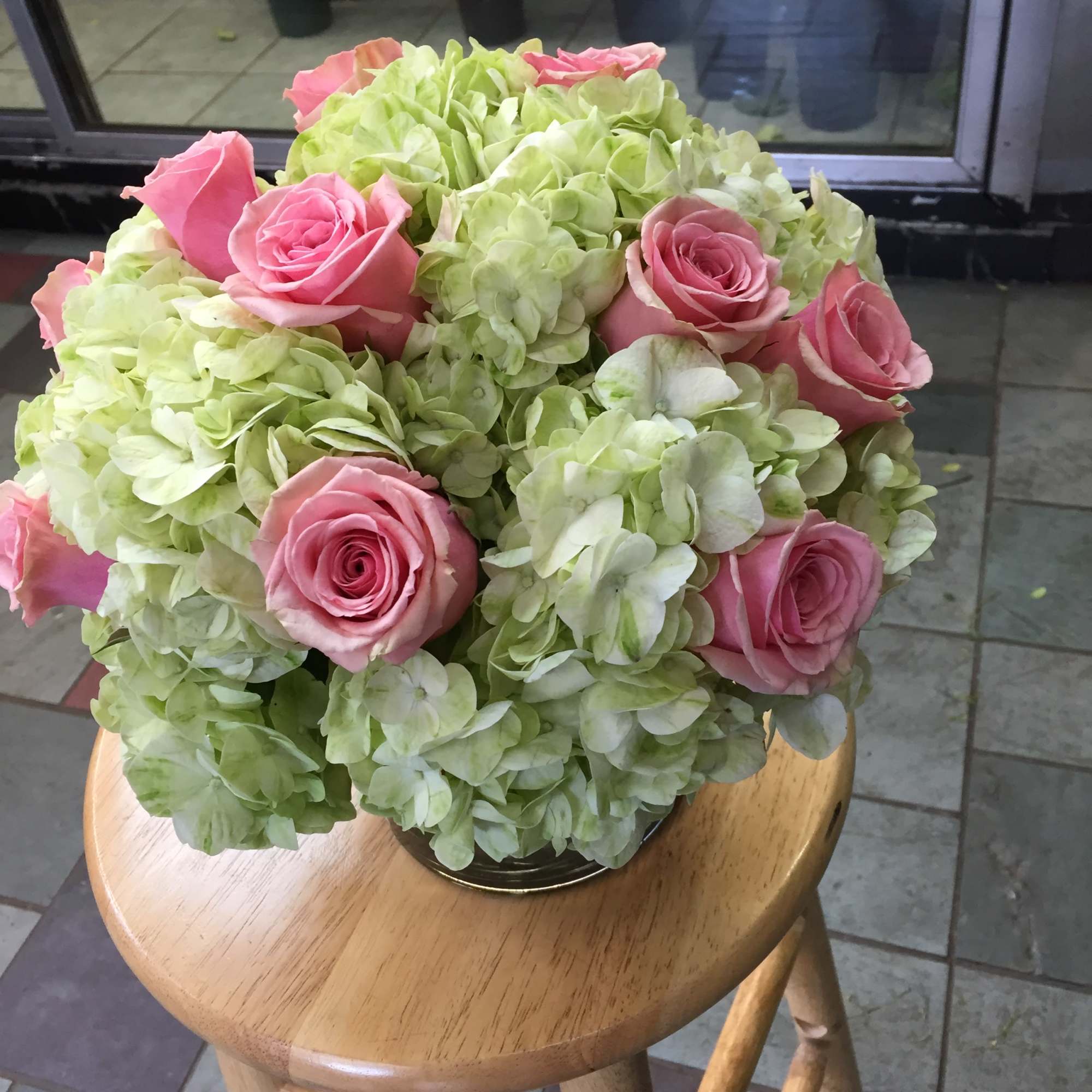 Round arrangement of pale green hydrangeas and pink roses in a clear glass vase on a wooden stool