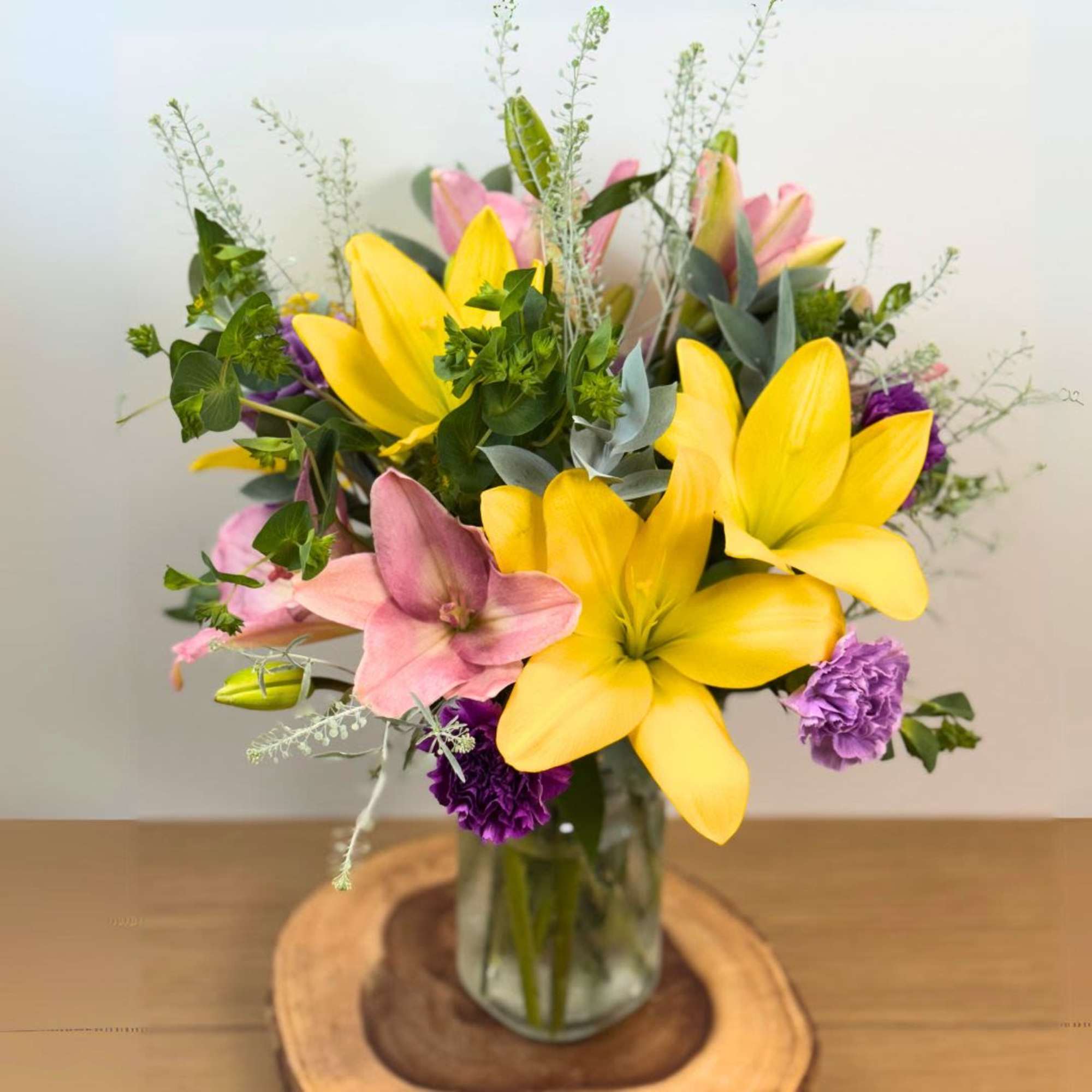 Arrangement of yellow lilies with pink lilies and purple carnations in a clear glass vase on a wooden base.