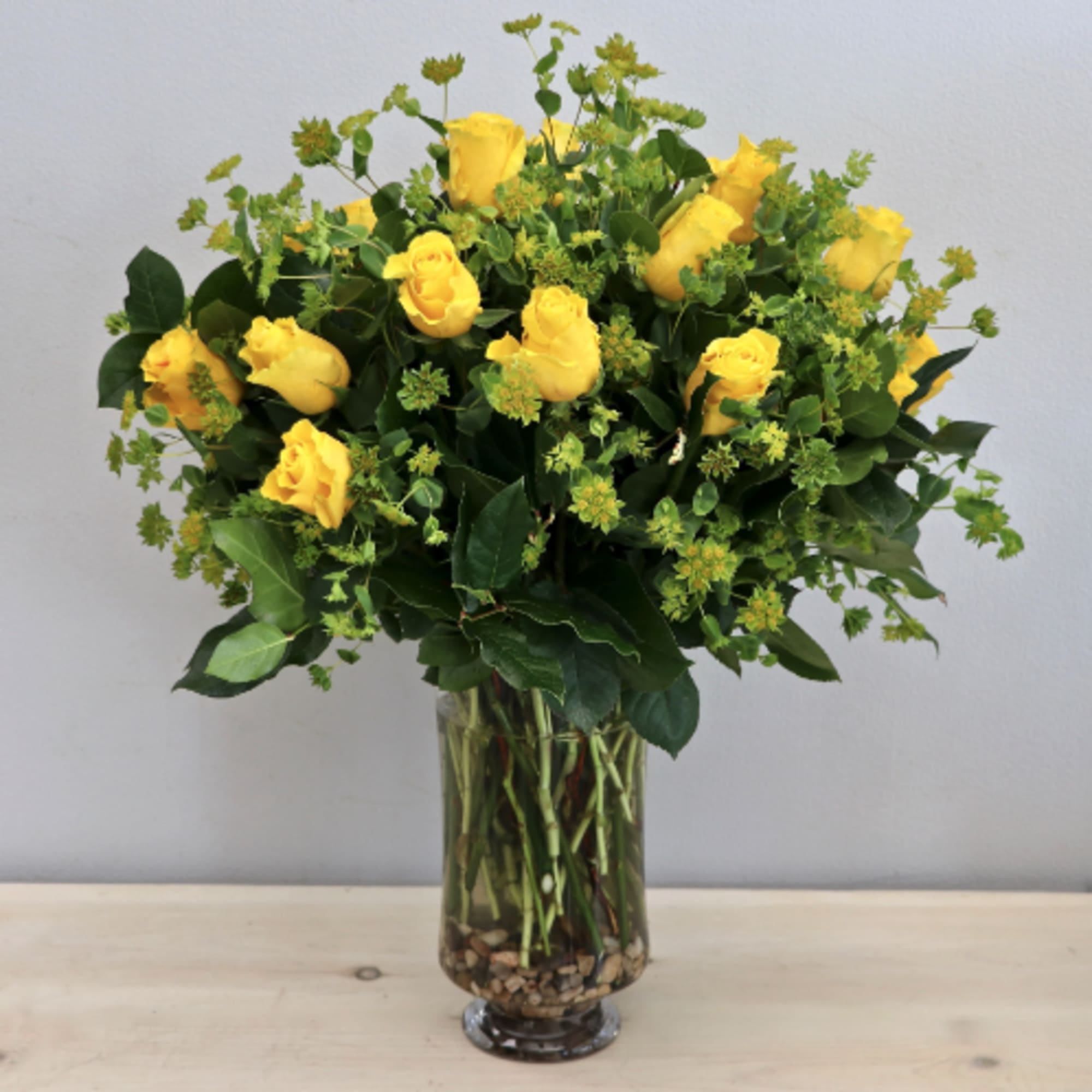 Tall arrangement of yellow roses with green filler in a clear glass vase with stones