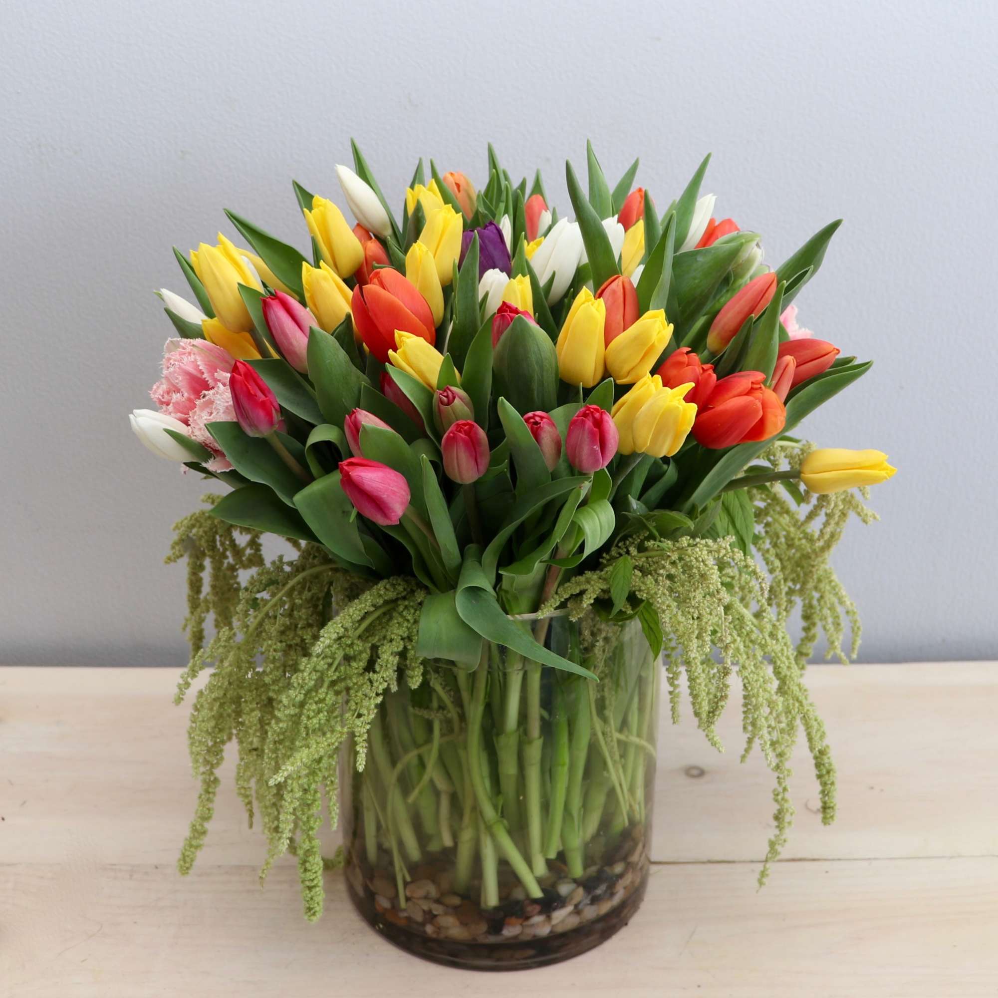Large mixed-color tulip arrangement with trailing green accents in a clear glass vase with stones