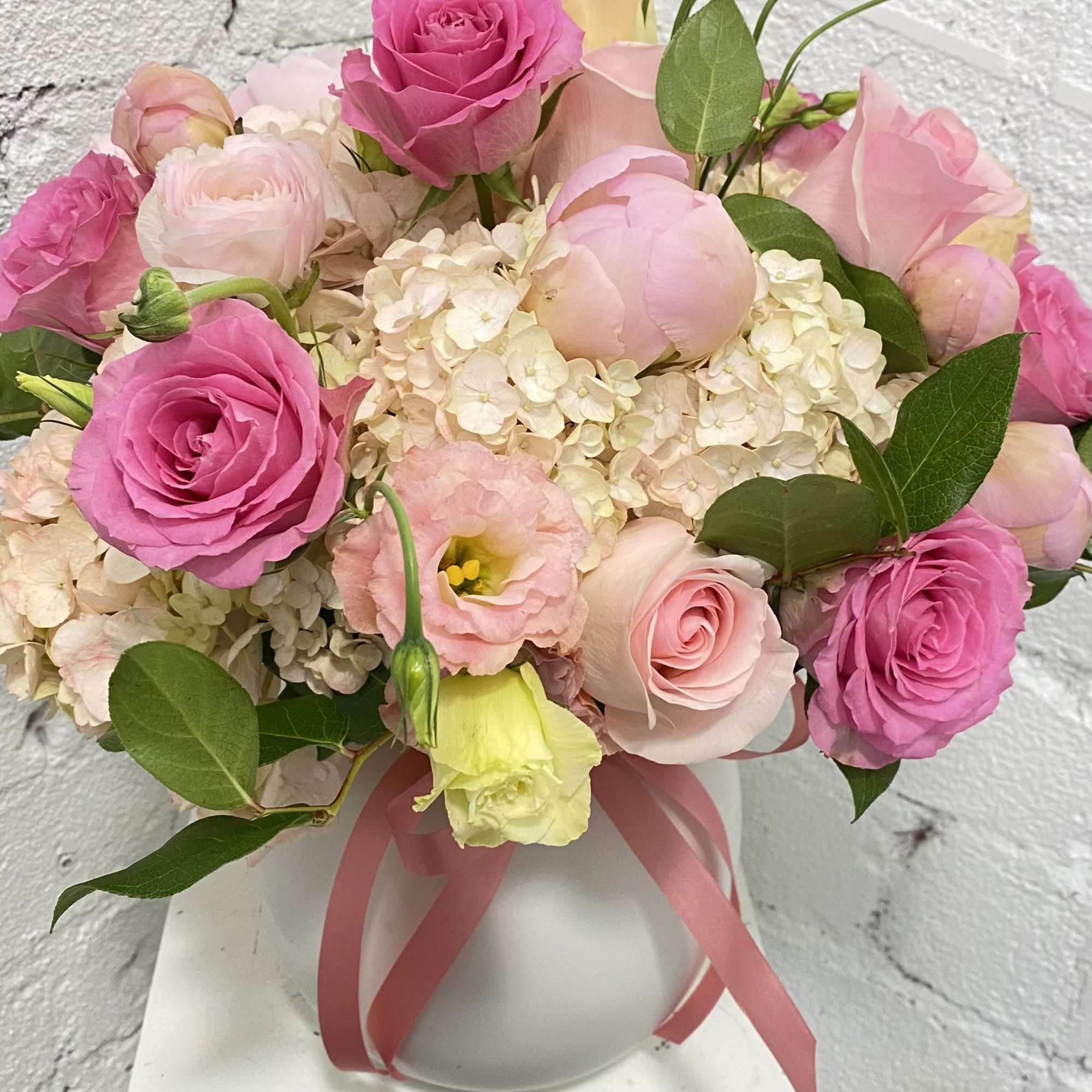 Pink roses and pale blooms arranged in a white vase with a ribbon