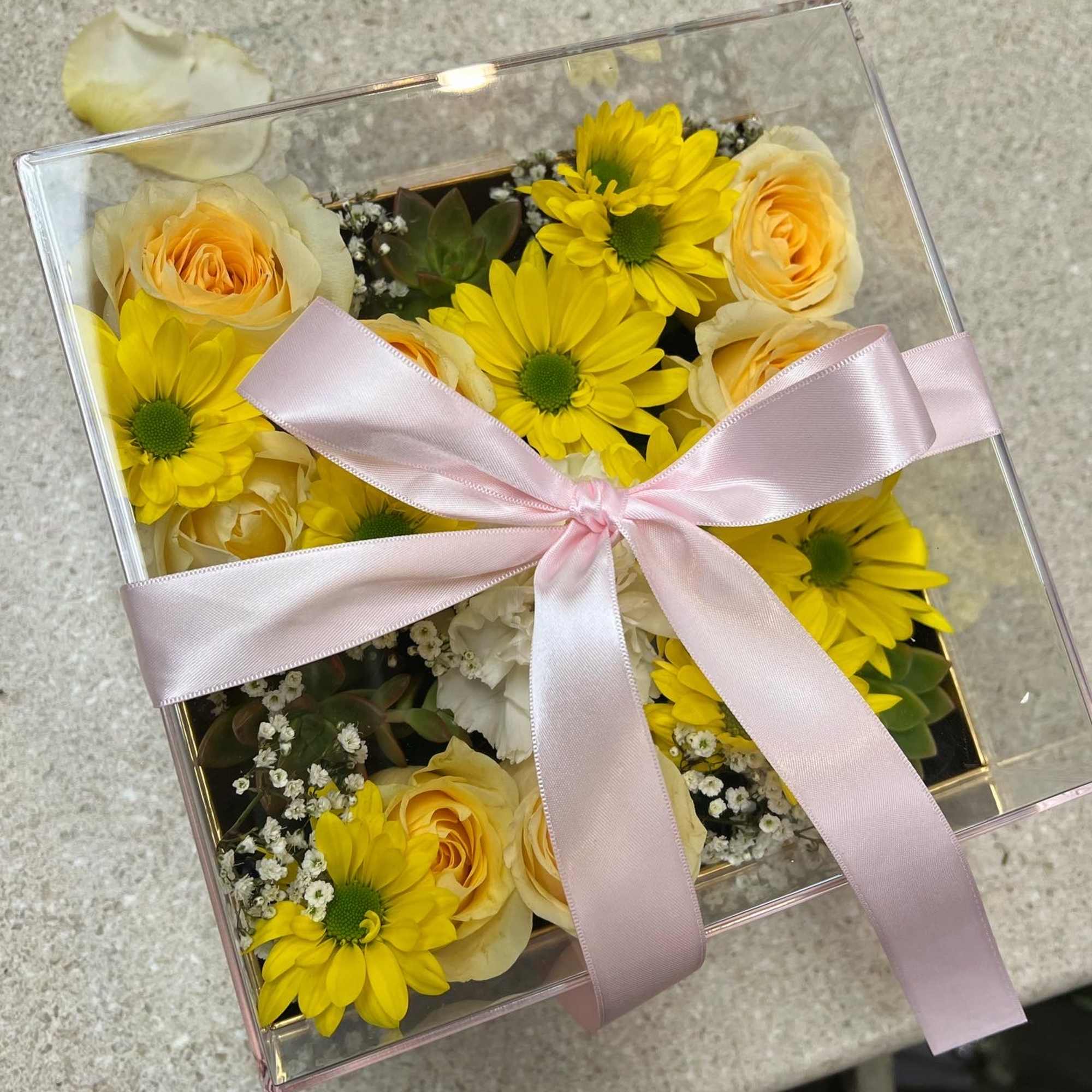 Yellow daisies and pale peach roses in a clear gift box with a pink ribbon.
