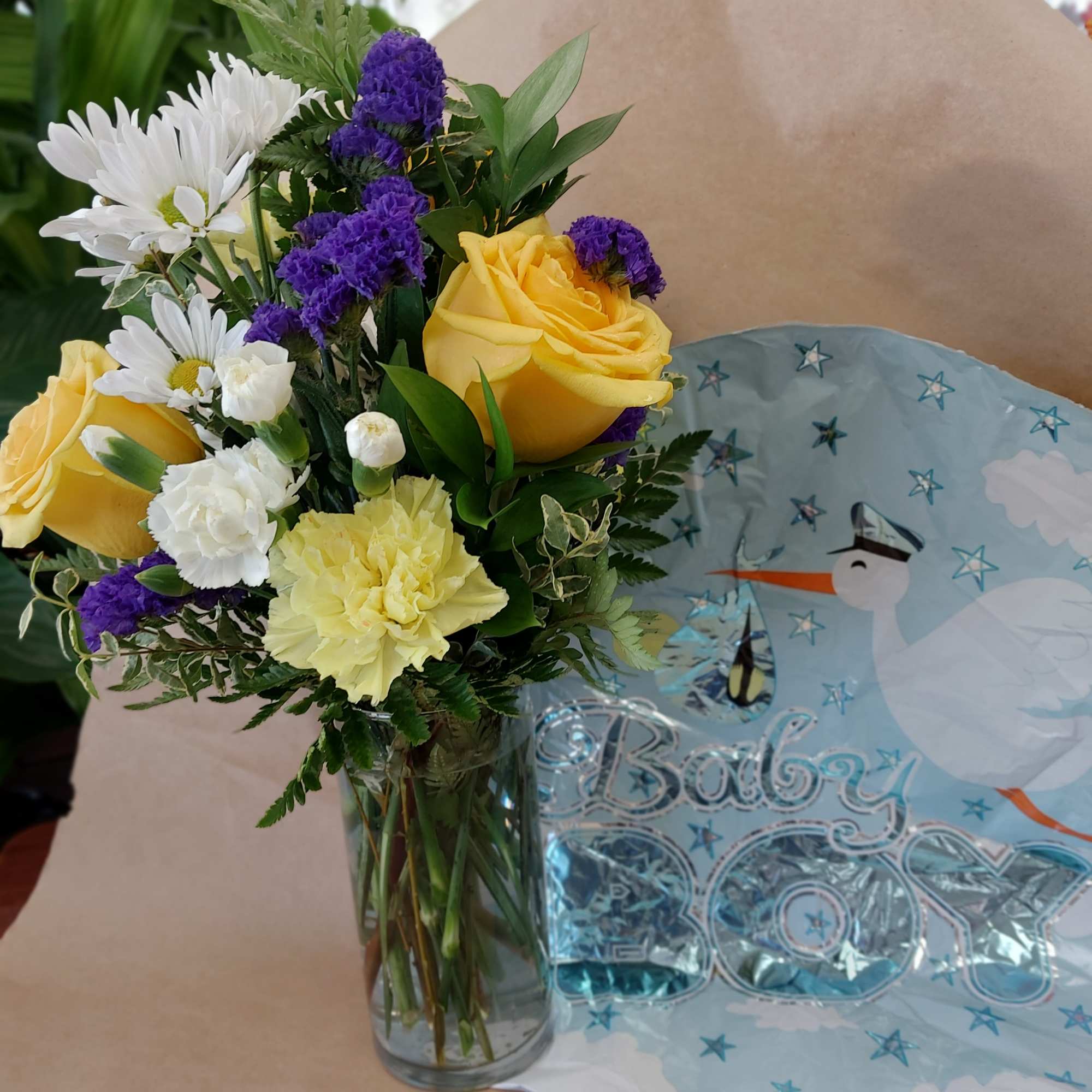 Bouquet of yellow roses, white daisies, and purple filler in a glass vase beside a baby balloon