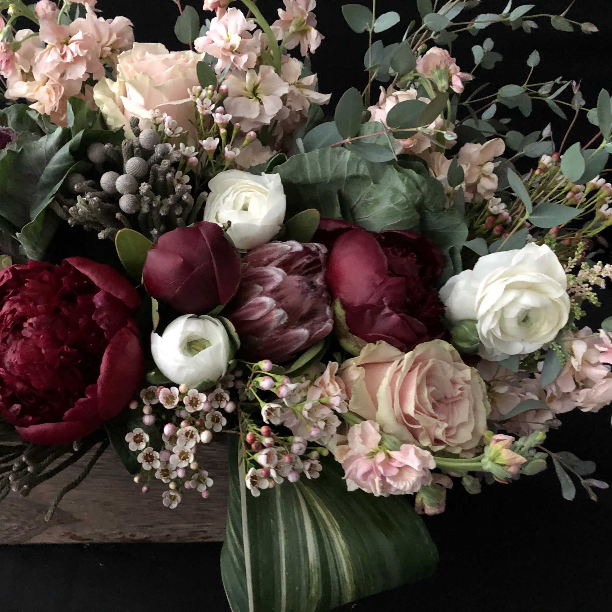 Low arrangement of burgundy peonies, blush roses, and white ranunculus in a wooden box