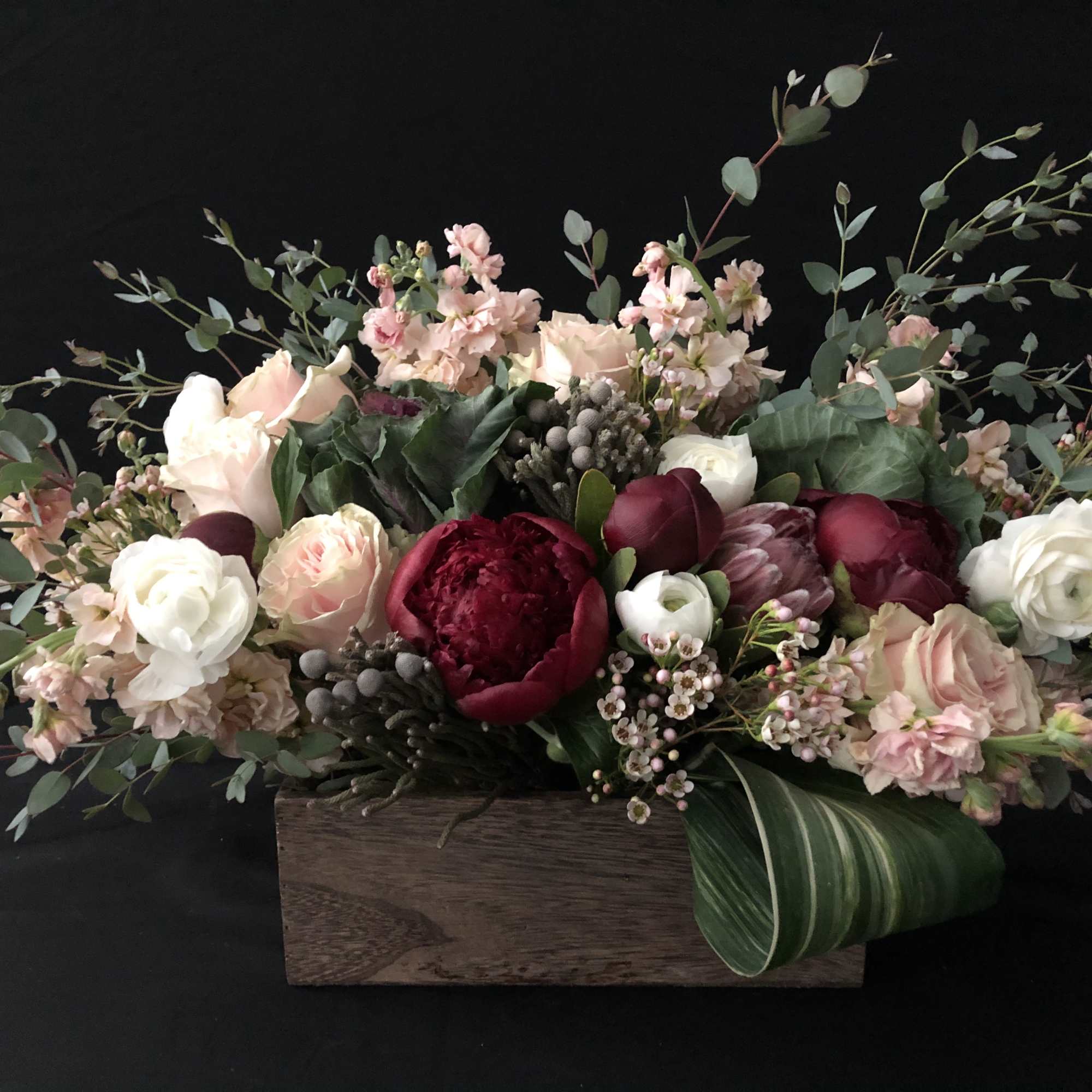 Low arrangement of blush and white flowers with deep red blooms in a wooden box with a large green leaf.