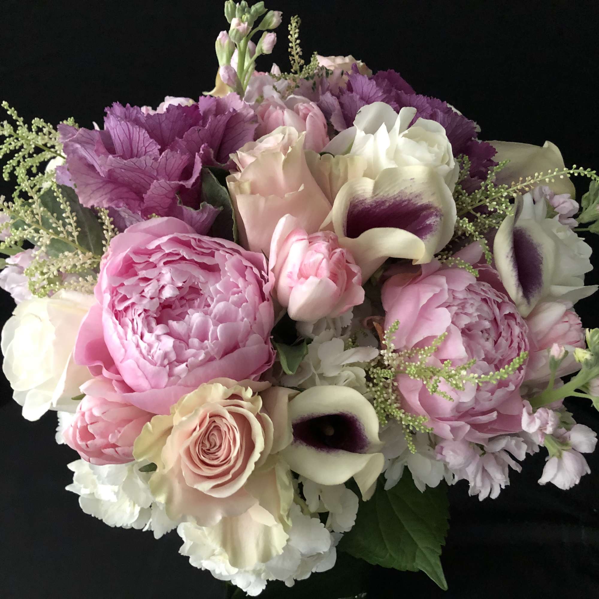 Round bouquet of pink peonies, roses, and white mini calla lilies with purple centers on a dark background