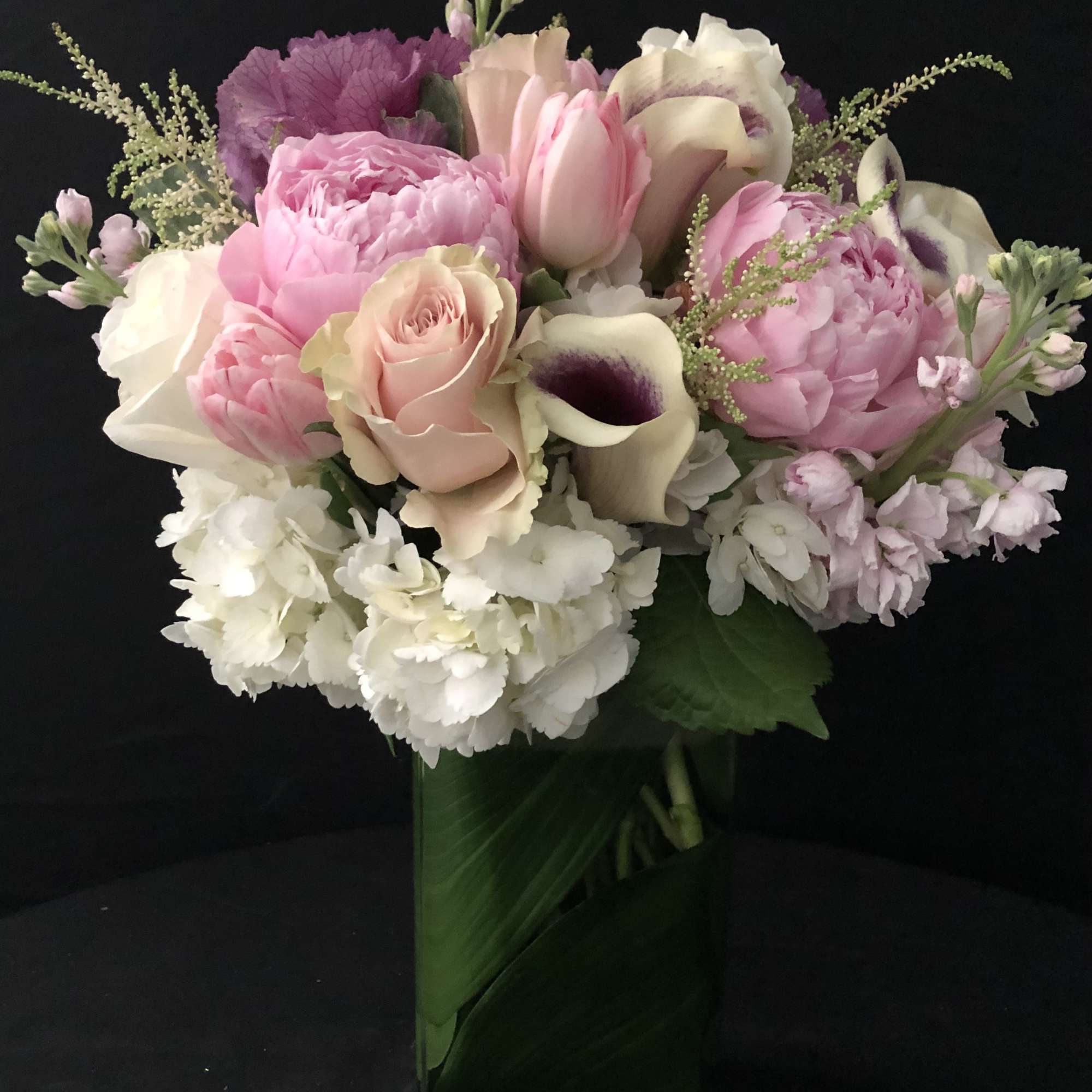 Arrangement of pink peonies, roses, tulips, and white hydrangeas in a tall glass vase lined with green leaves