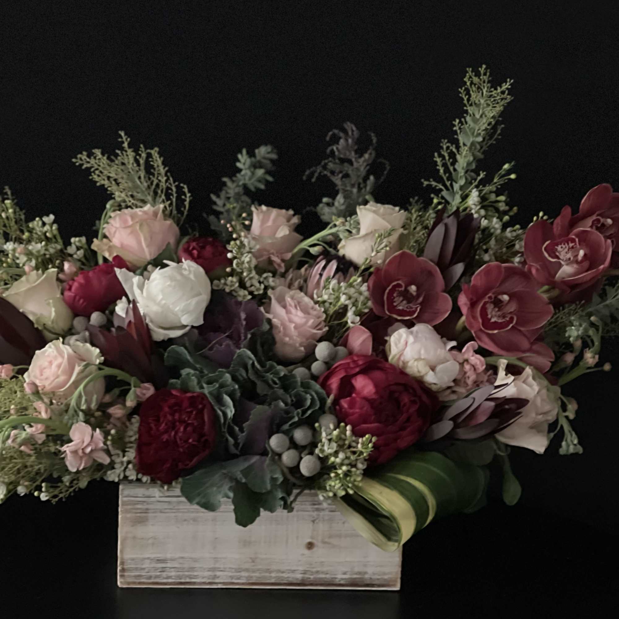 Low wooden box arrangement with red orchids, blush and white roses, peonies, and looped green leaves.