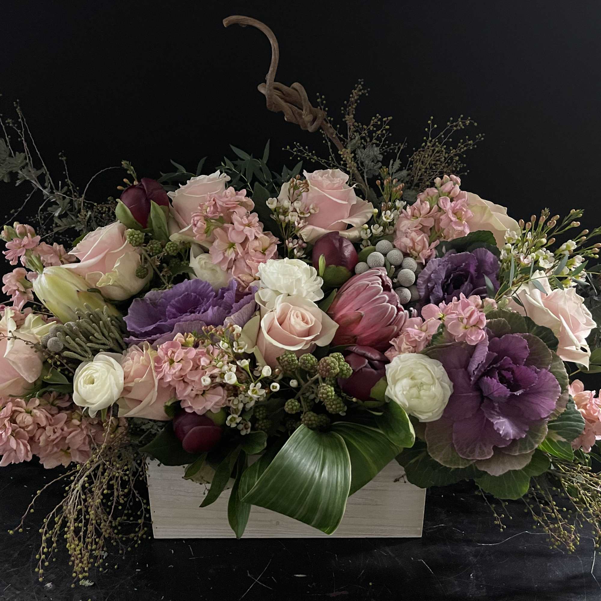 Low rectangular arrangement of pink roses, purple blooms, and white flowers in a wooden box.