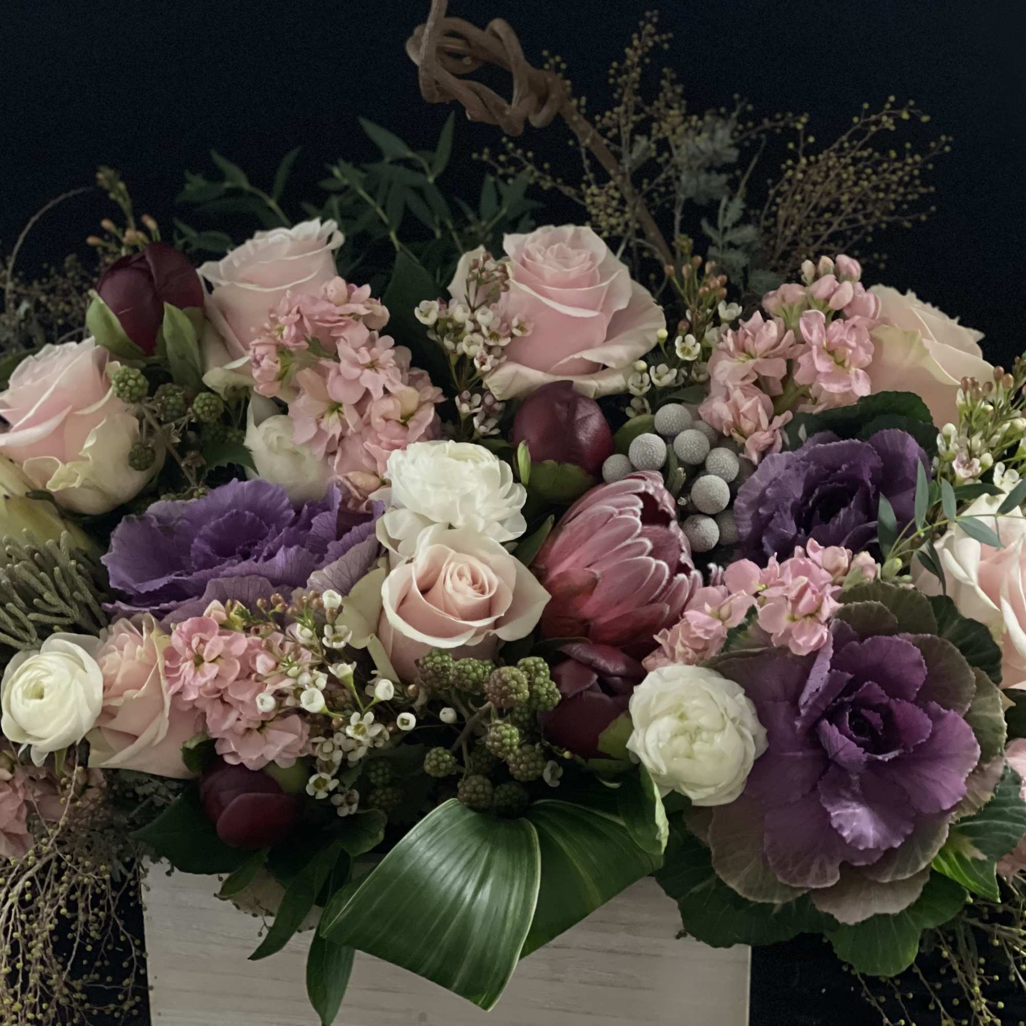 Low pink and purple arrangement with roses, protea, tulips and ornamental cabbage in a white wooden box