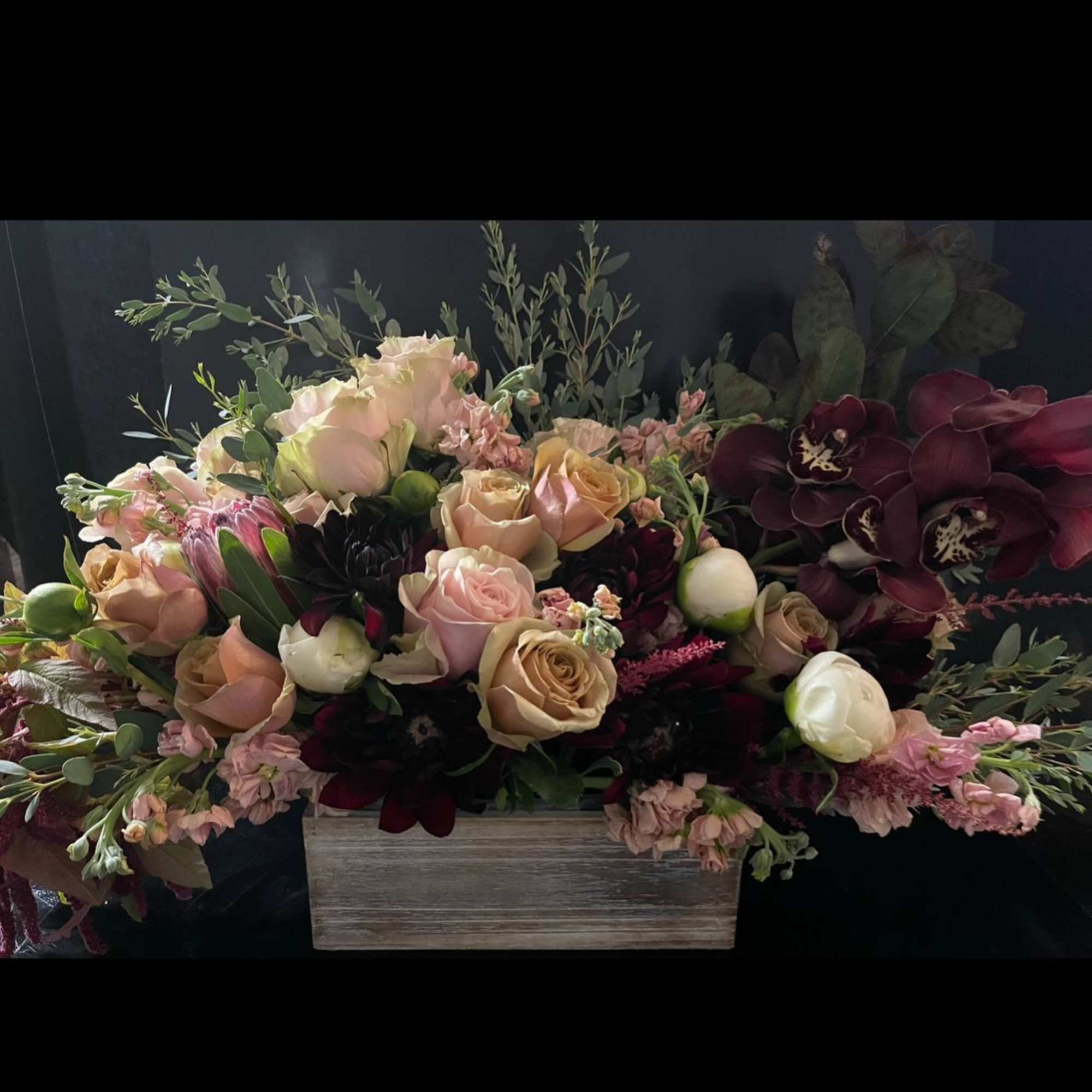 Low centerpiece of blush and peach roses with dark burgundy blooms in a rustic wooden box.