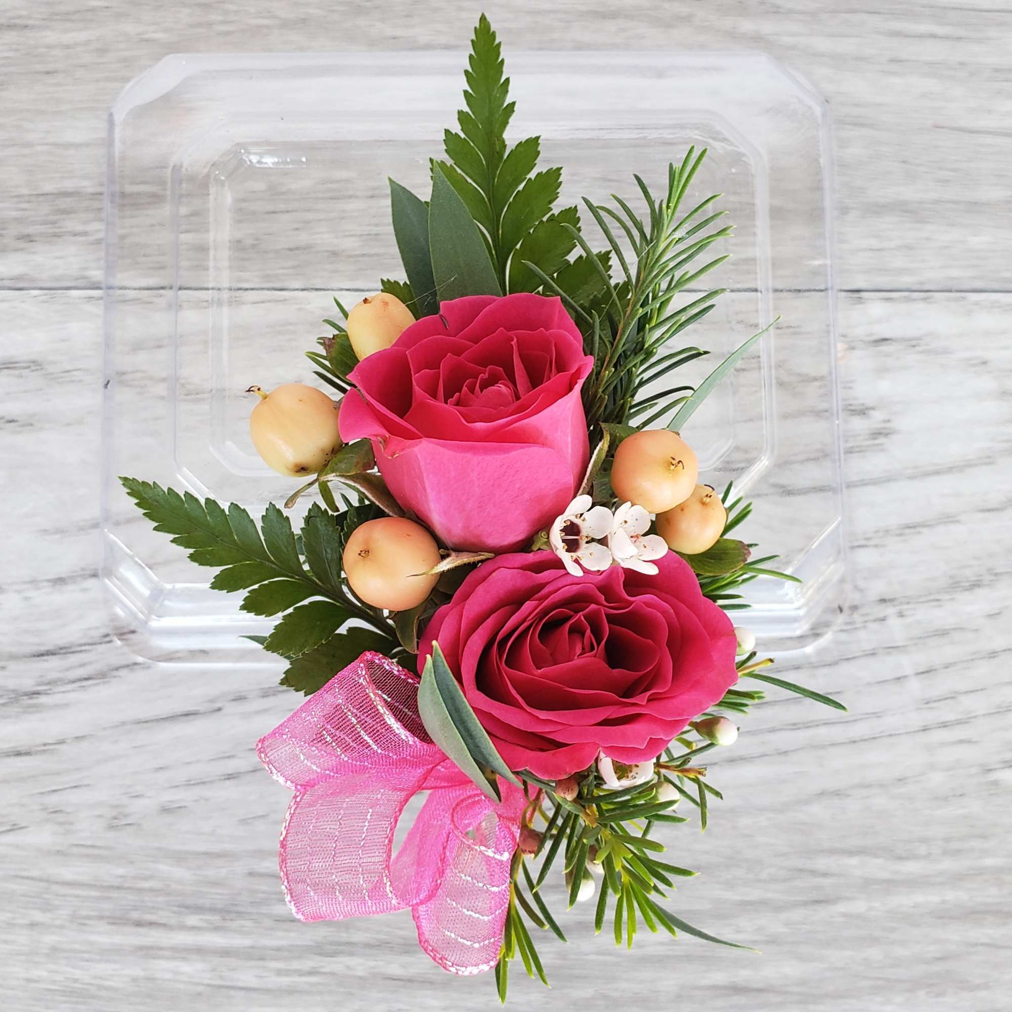 Two pink roses with berries and a pink ribbon in a clear plastic box