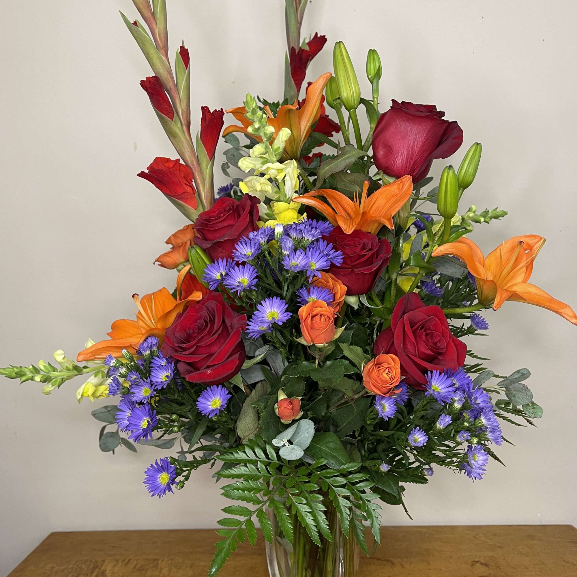 Mixed bouquet of red roses, orange lilies, and purple daisies in a clear vase