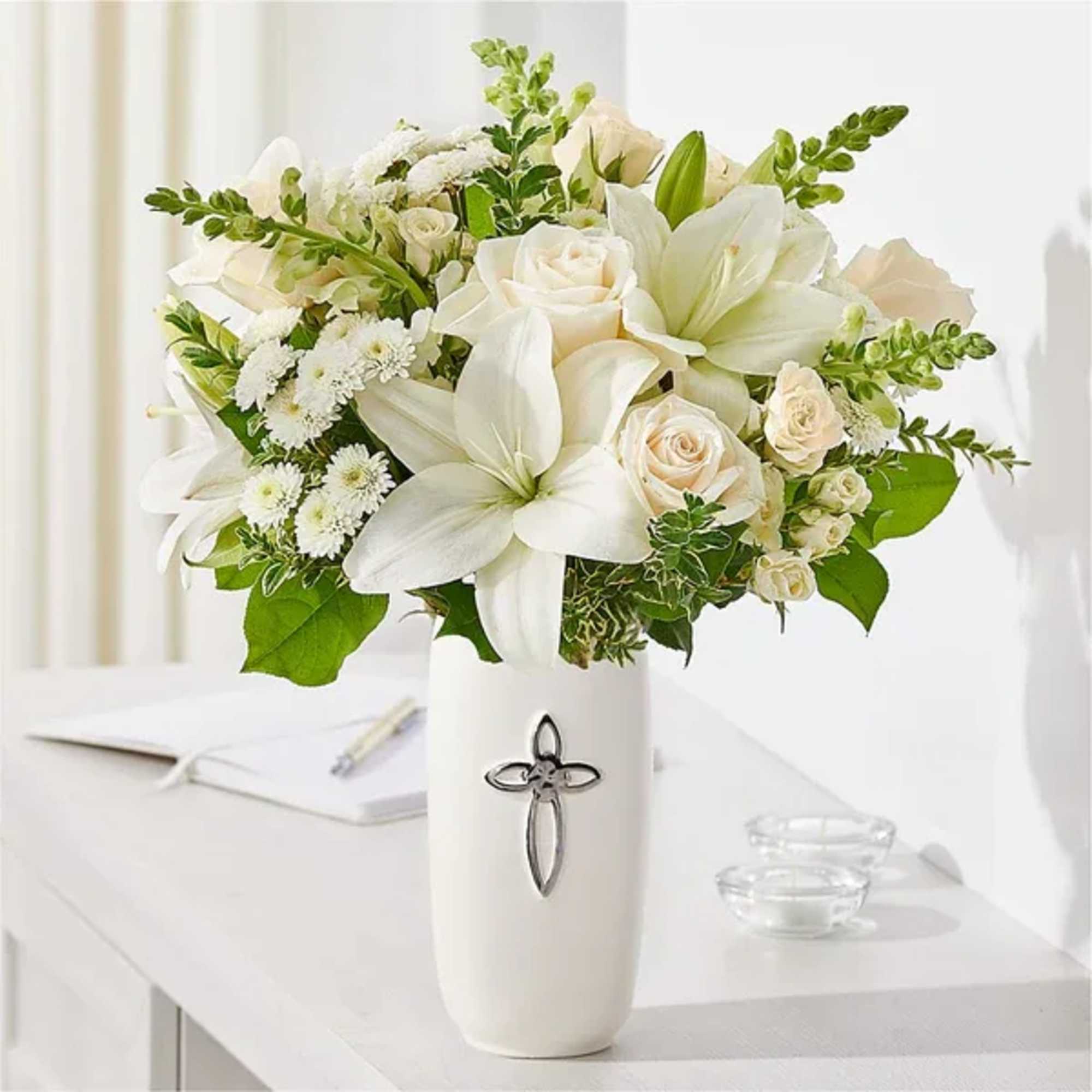 White lilies and roses arranged in a tall white vase with a cross emblem