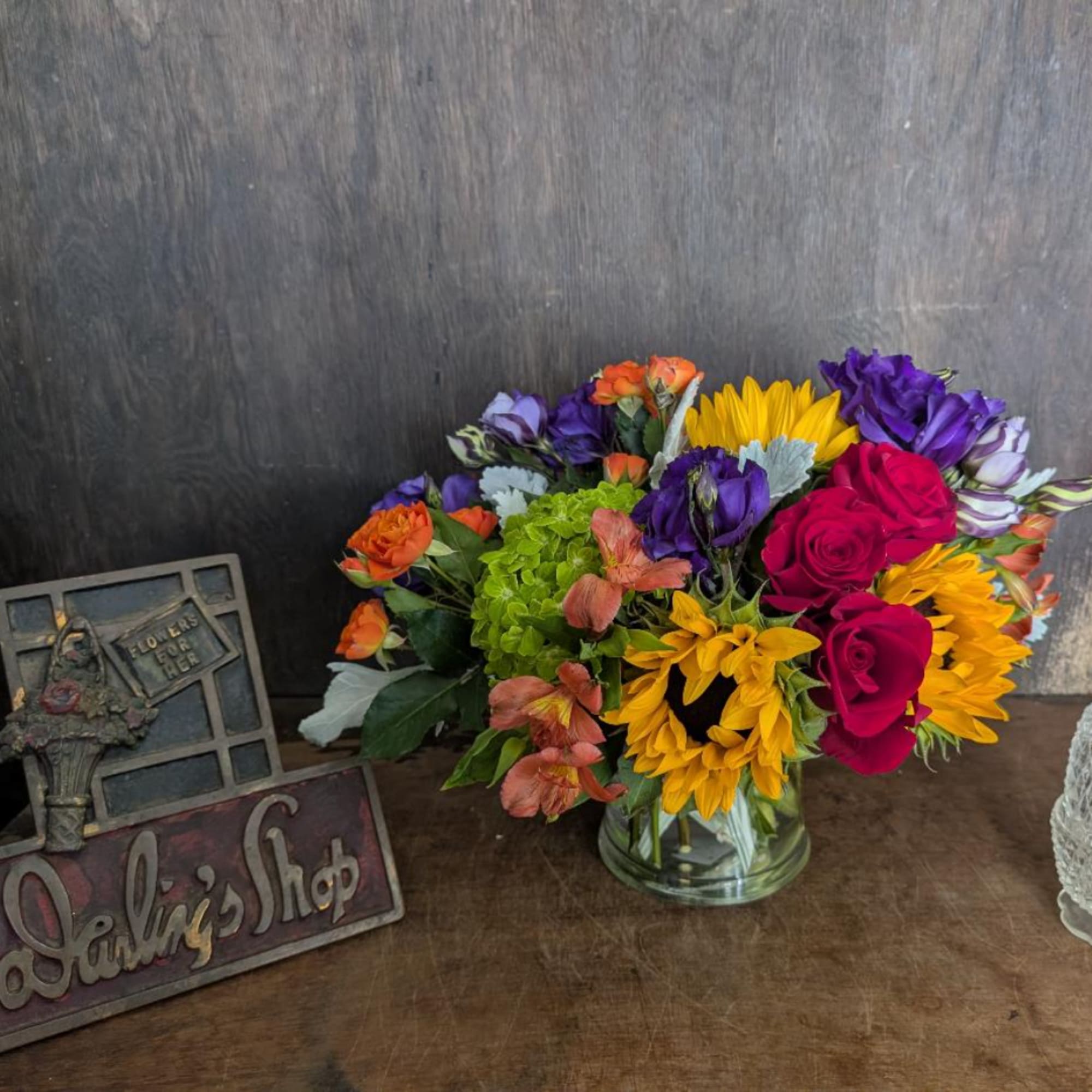 Mixed bouquet in a clear glass vase with roses, sunflowers, and purple blooms