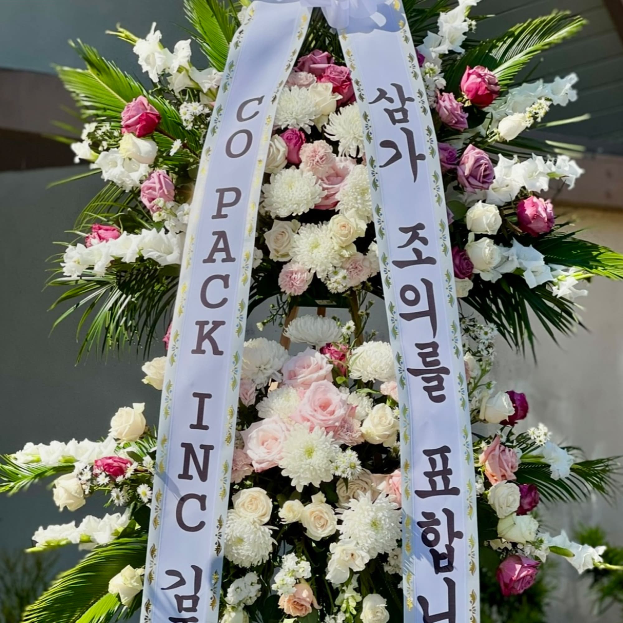 Tall standing floral wreath with pink and white flowers and two white condolence ribbons