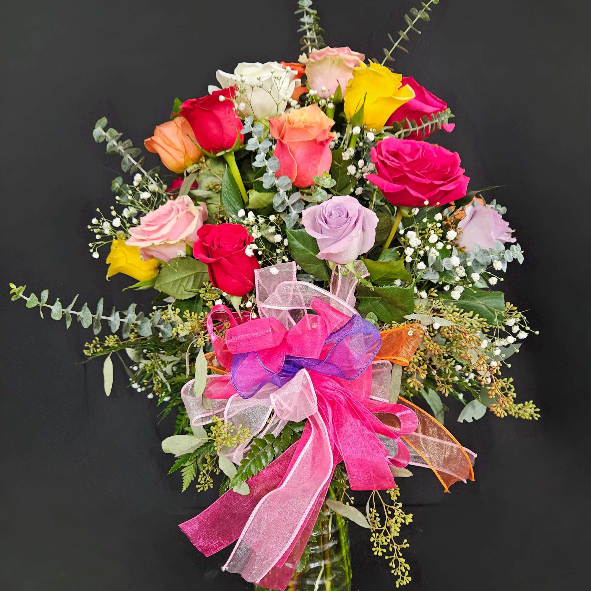Tall mixed-color rose arrangement in a clear glass vase with a large pink and purple ribbon bow.