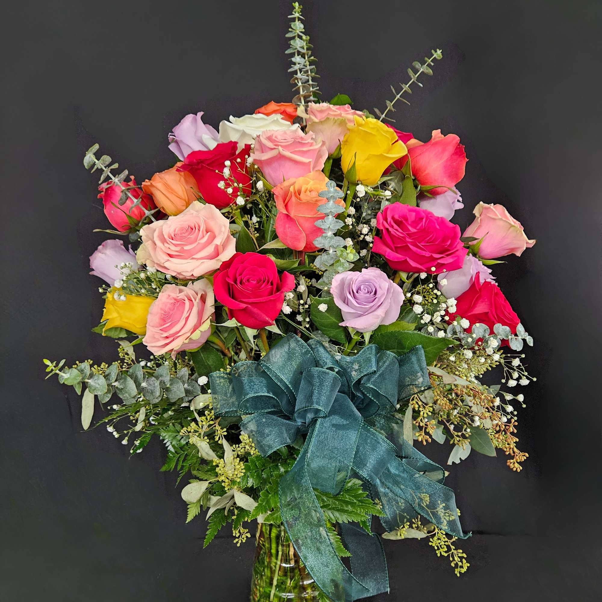 Tall arrangement of multicolored roses with baby's breath in a clear vase with a large teal bow.