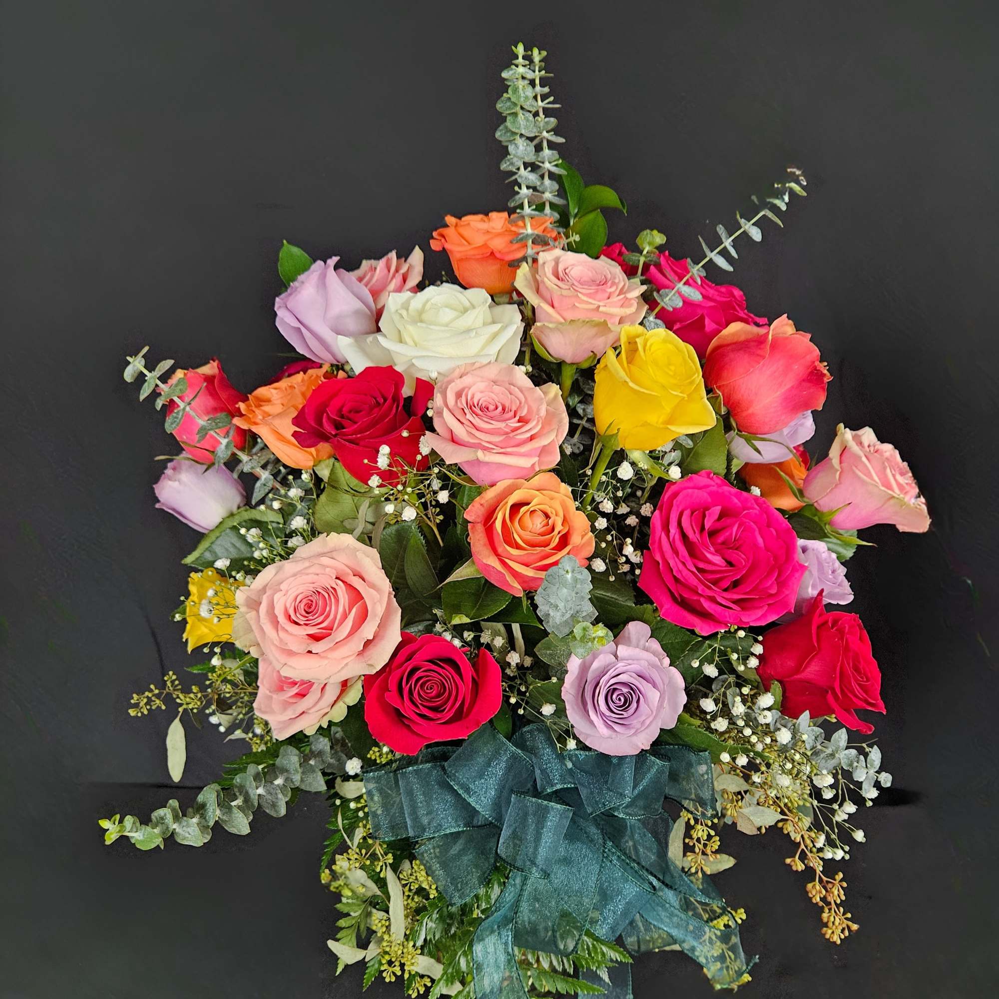 Arrangement of multicolored roses with filler flowers and a green ribbon bow on a dark background