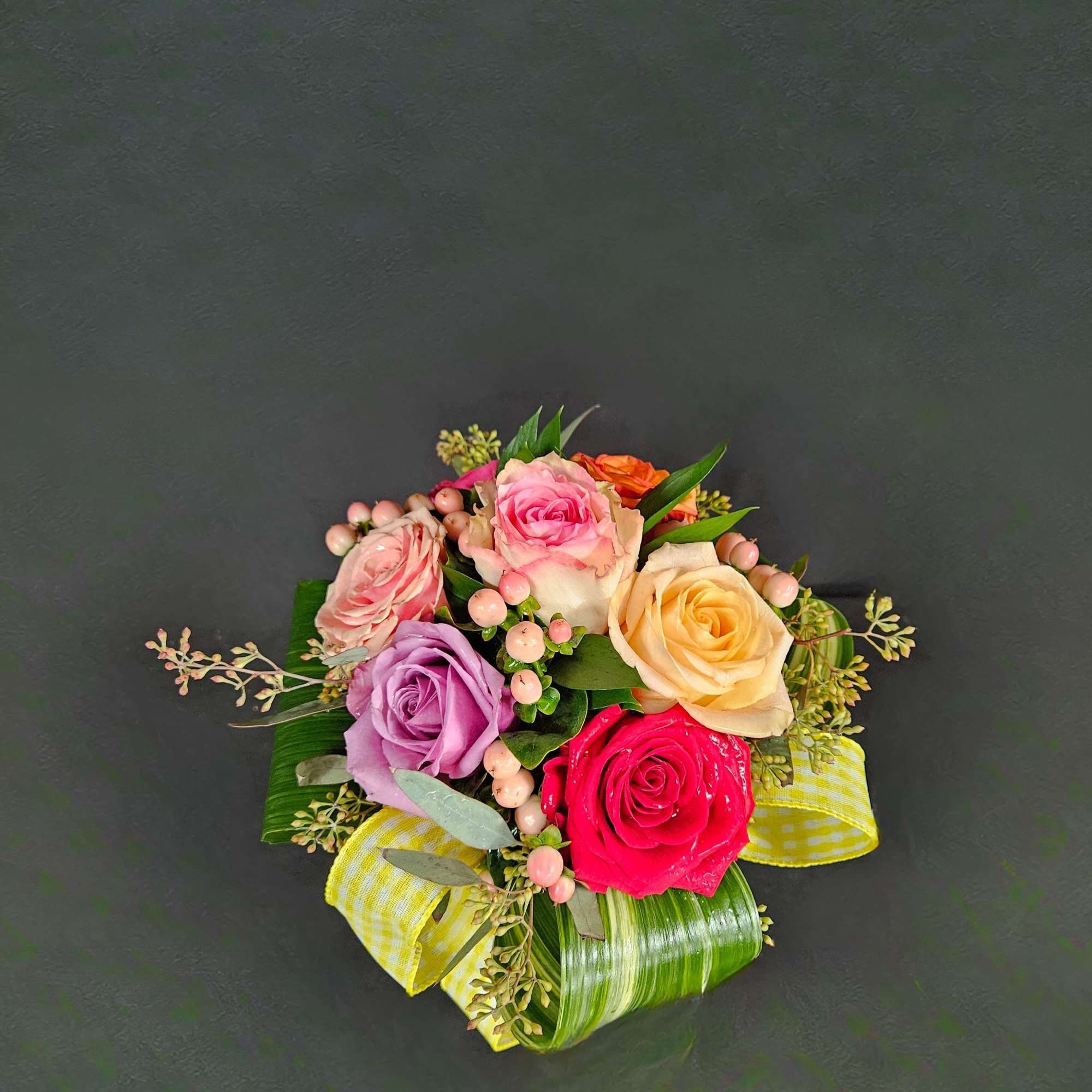 Small round bouquet of mixed-color roses with pink berries and yellow checked ribbon on a dark background