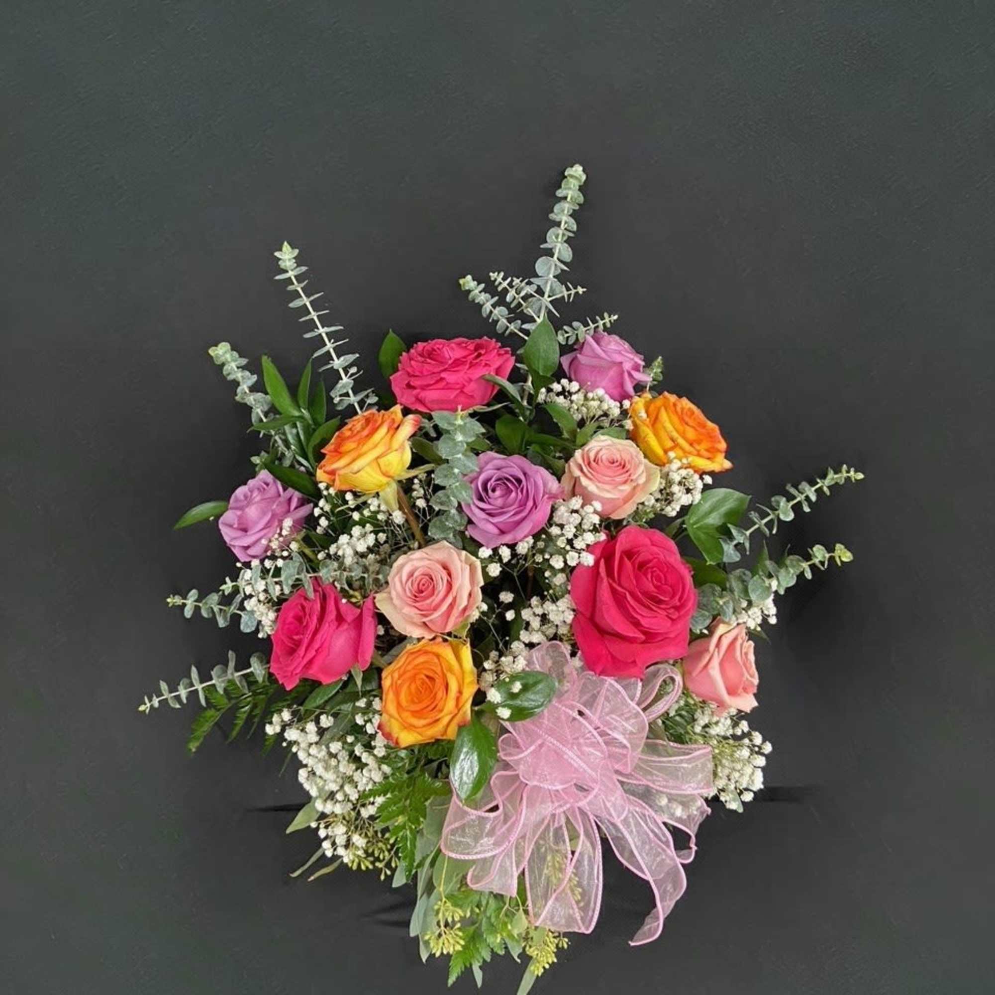 Round arrangement of mixed pink, lavender, and orange roses with baby's breath and a large pink bow.